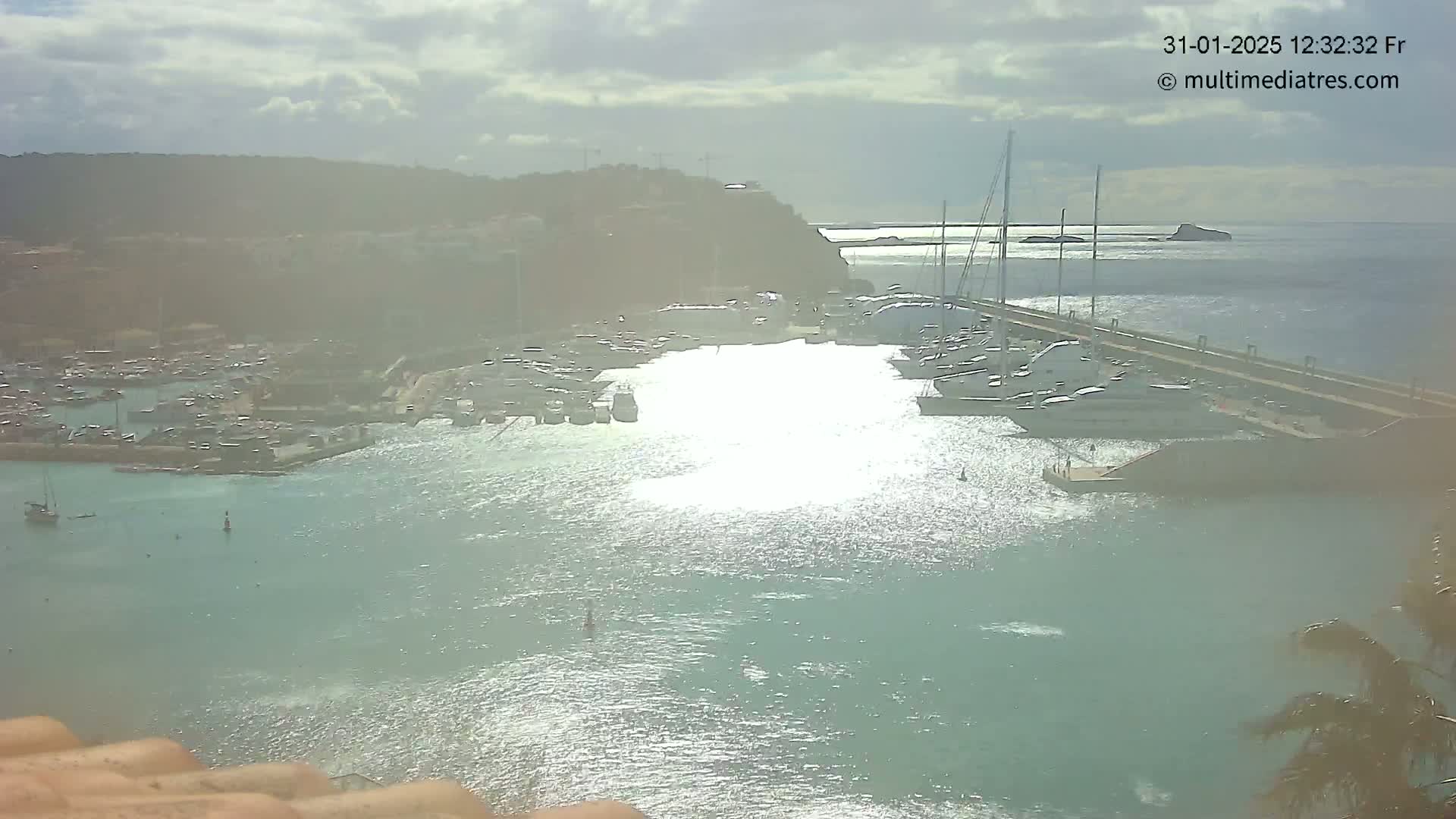 A harbor filled with numerous boats and yachts is seen under a partly cloudy sky, with a small island visible in the distance.