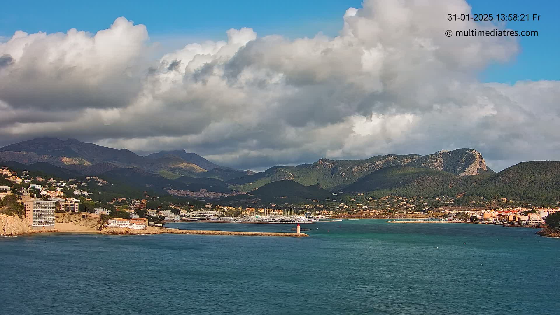 A coastal town nestled at the base of a mountain range under a partly cloudy sky, with a marina visible in the foreground.