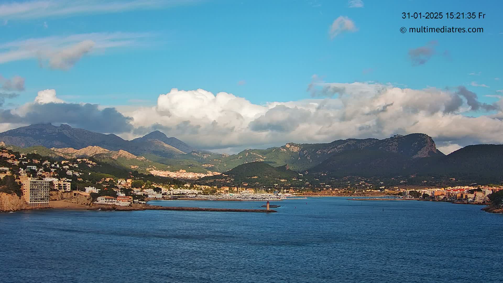 A coastal town nestled at the base of a mountain range under a partly cloudy sky sits on a calm body of water.