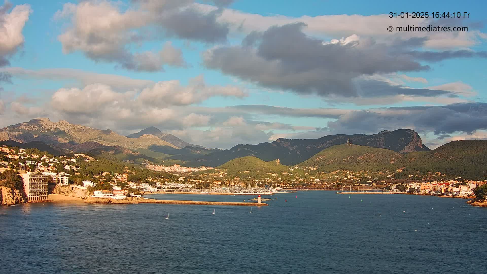 A partly cloudy sky overlooks a coastal town nestled between mountains and a calm harbor with boats, under a partly cloudy sky.
