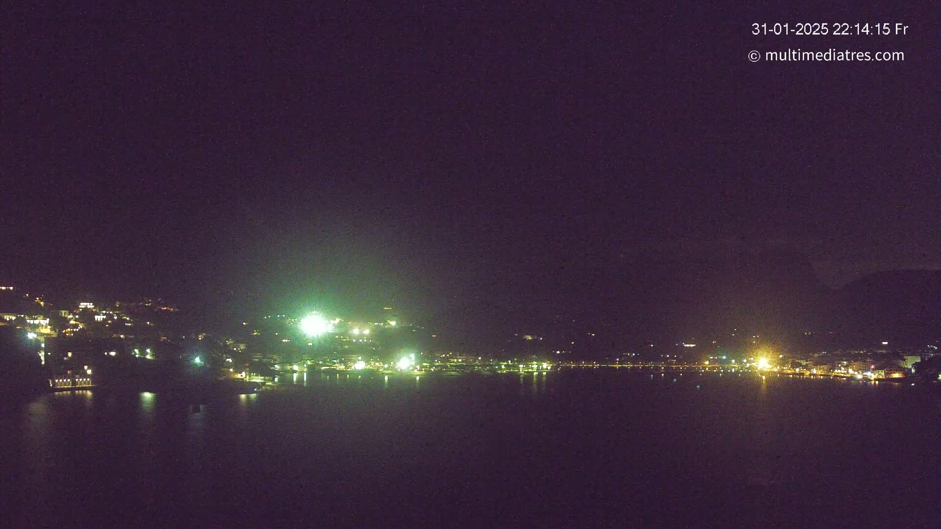 A nighttime view of a town situated on a body of water, with scattered lights illuminating the waterfront and hillside.