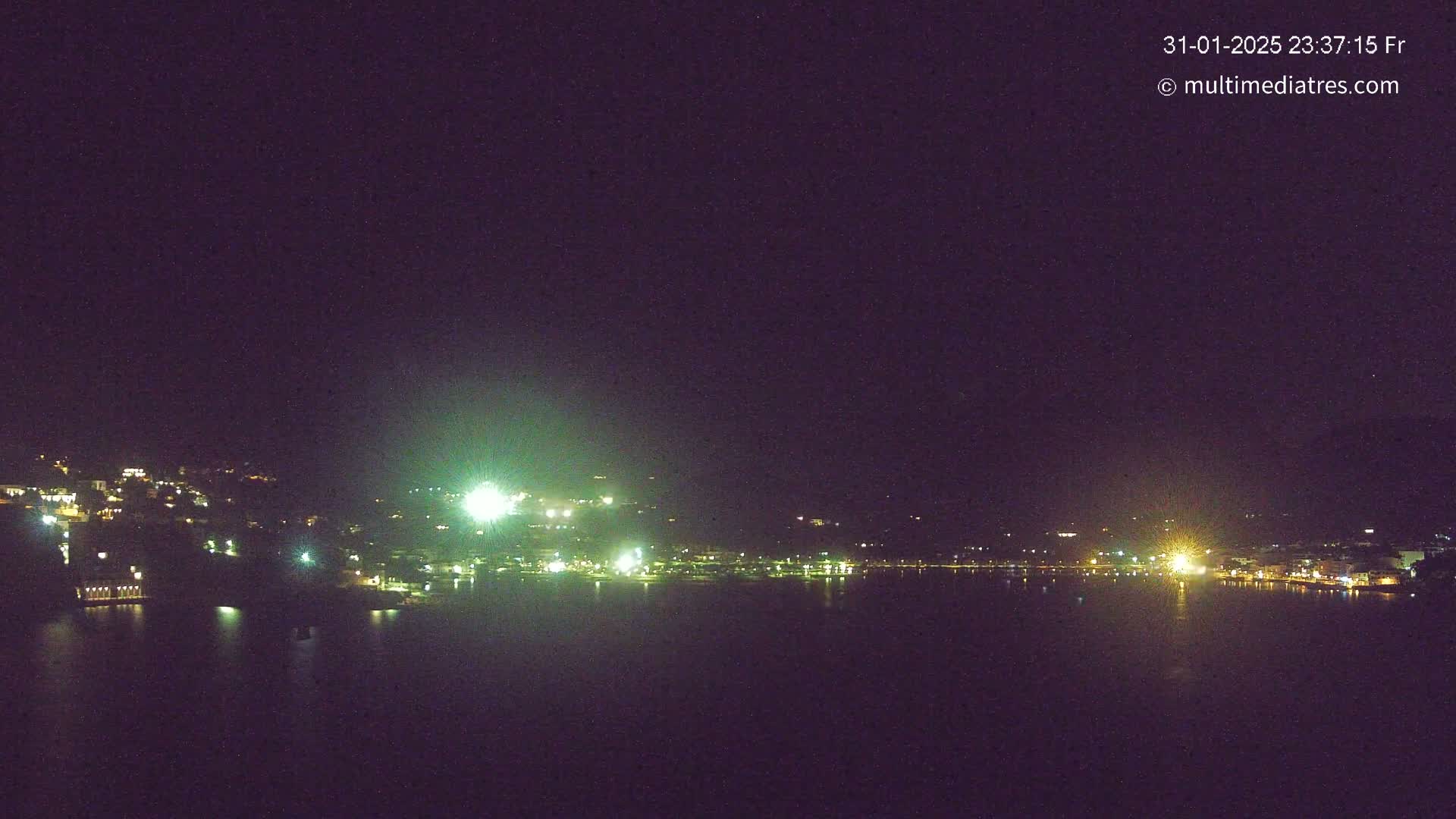 A nighttime long shot of a city on a hill overlooking a dark body of water under a clear, dark sky.
