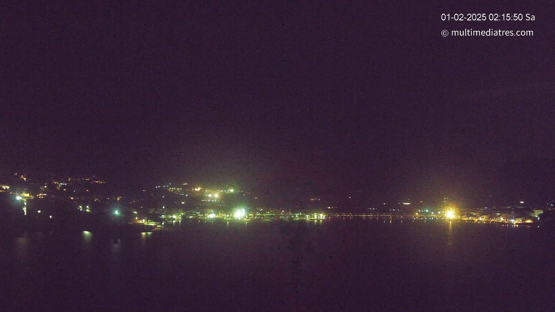 A nighttime view of a town's lights along a dark body of water under a clear, dark sky.