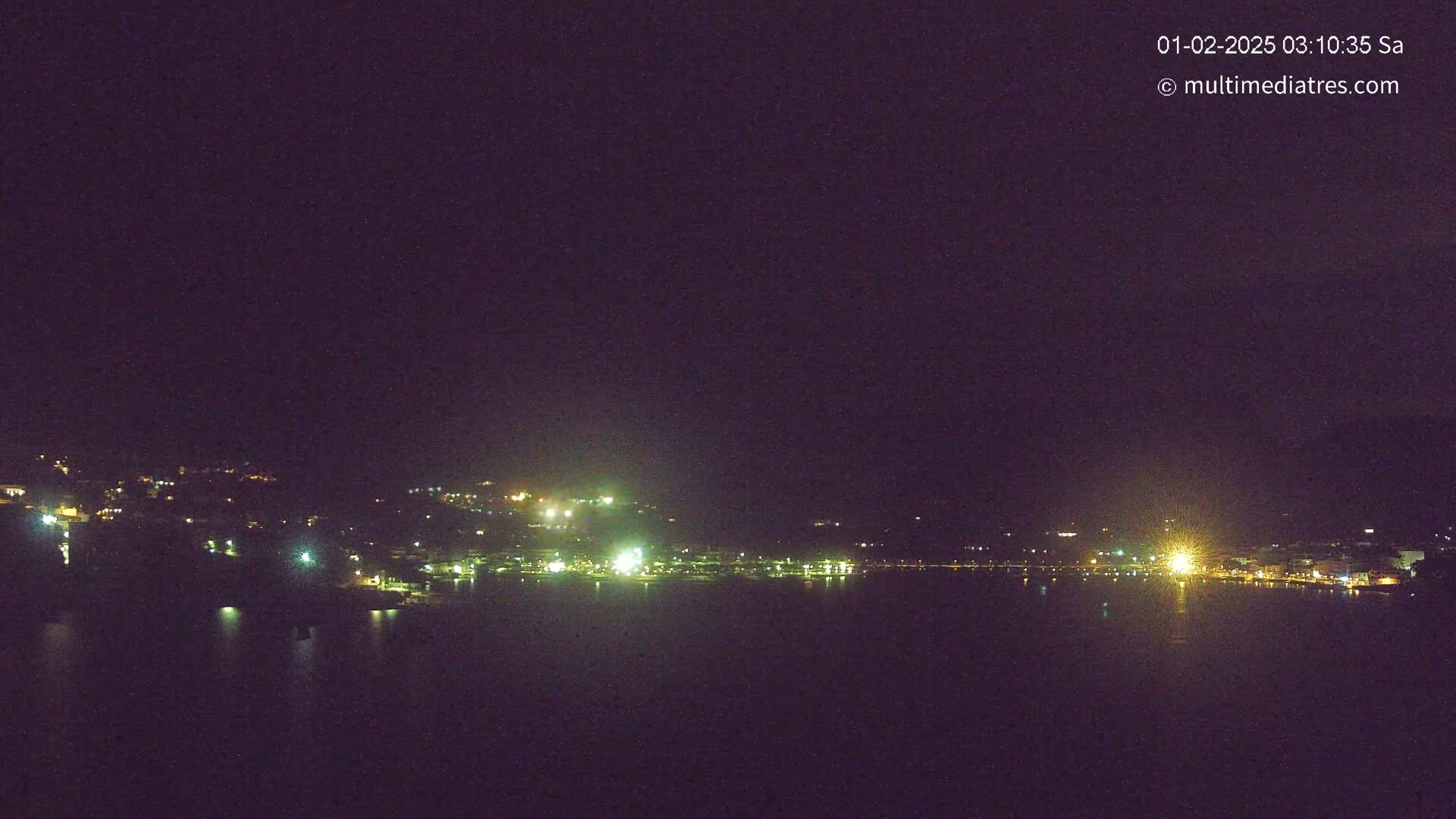 A nighttime, long shot of a town situated on a body of water, illuminated by scattered lights.