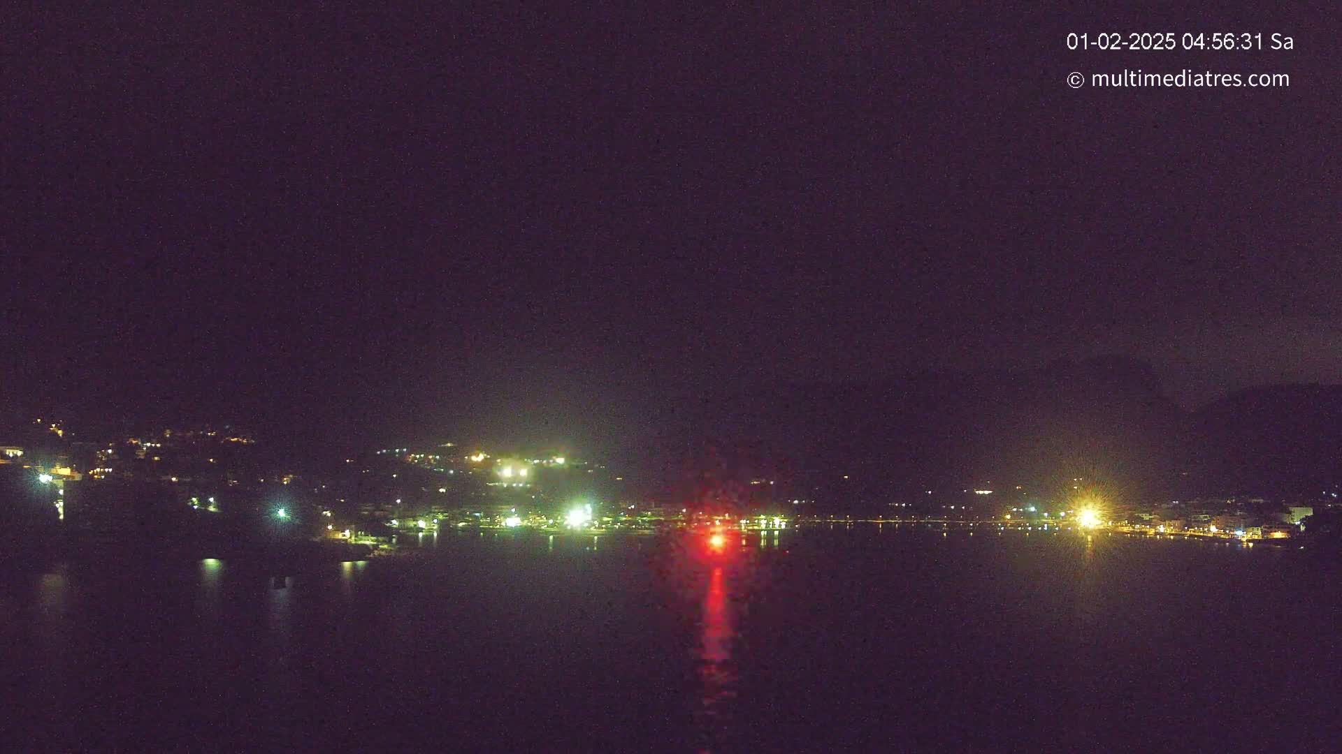A nighttime long shot of a town's lights across a dark body of water, with a single, bright red light reflecting on the water in the center.