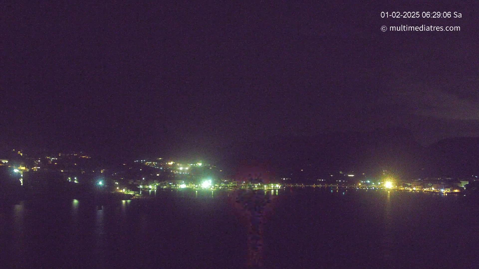 A nighttime, long-range view shows a dark body of water reflecting the lights of a town situated on a hillside.