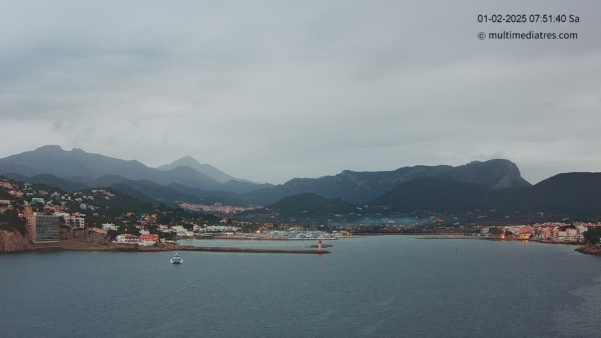 A coastal town nestled against a mountain range is viewed from across a calm bay under an overcast sky.
