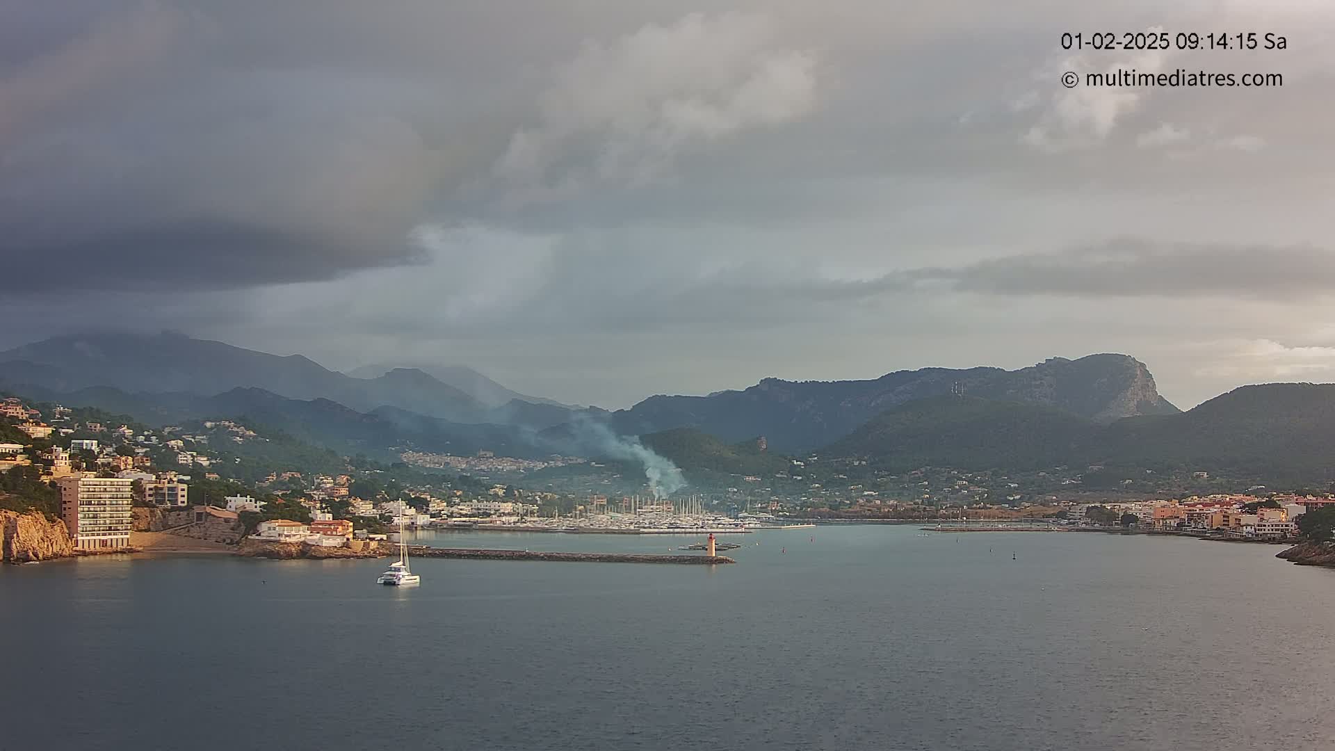 A coastal town nestled between mountains under a cloudy sky, with a marina filled with boats and a single sailboat on the water.