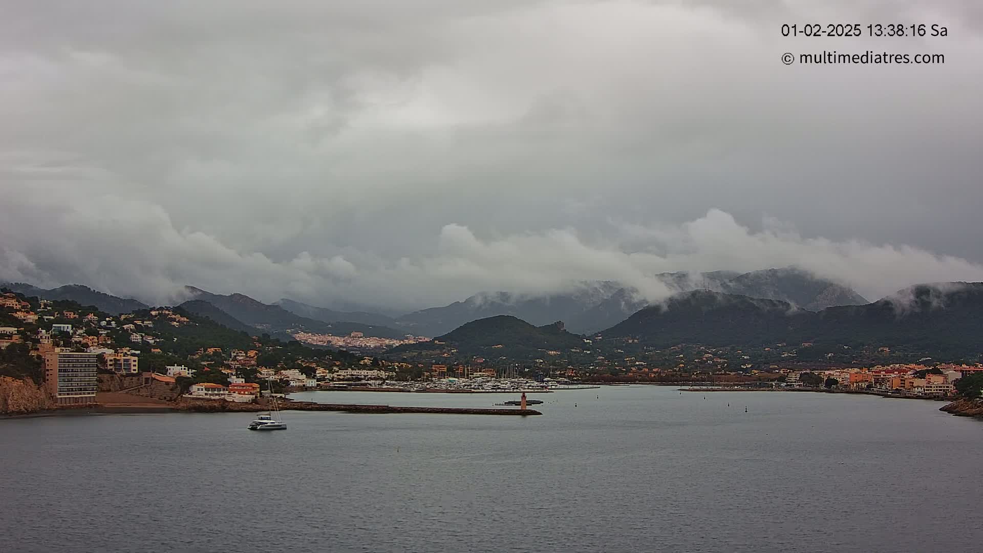A coastal town nestled against cloud-covered mountains is seen under an overcast sky, with a small boat on the calm water of a harbor.