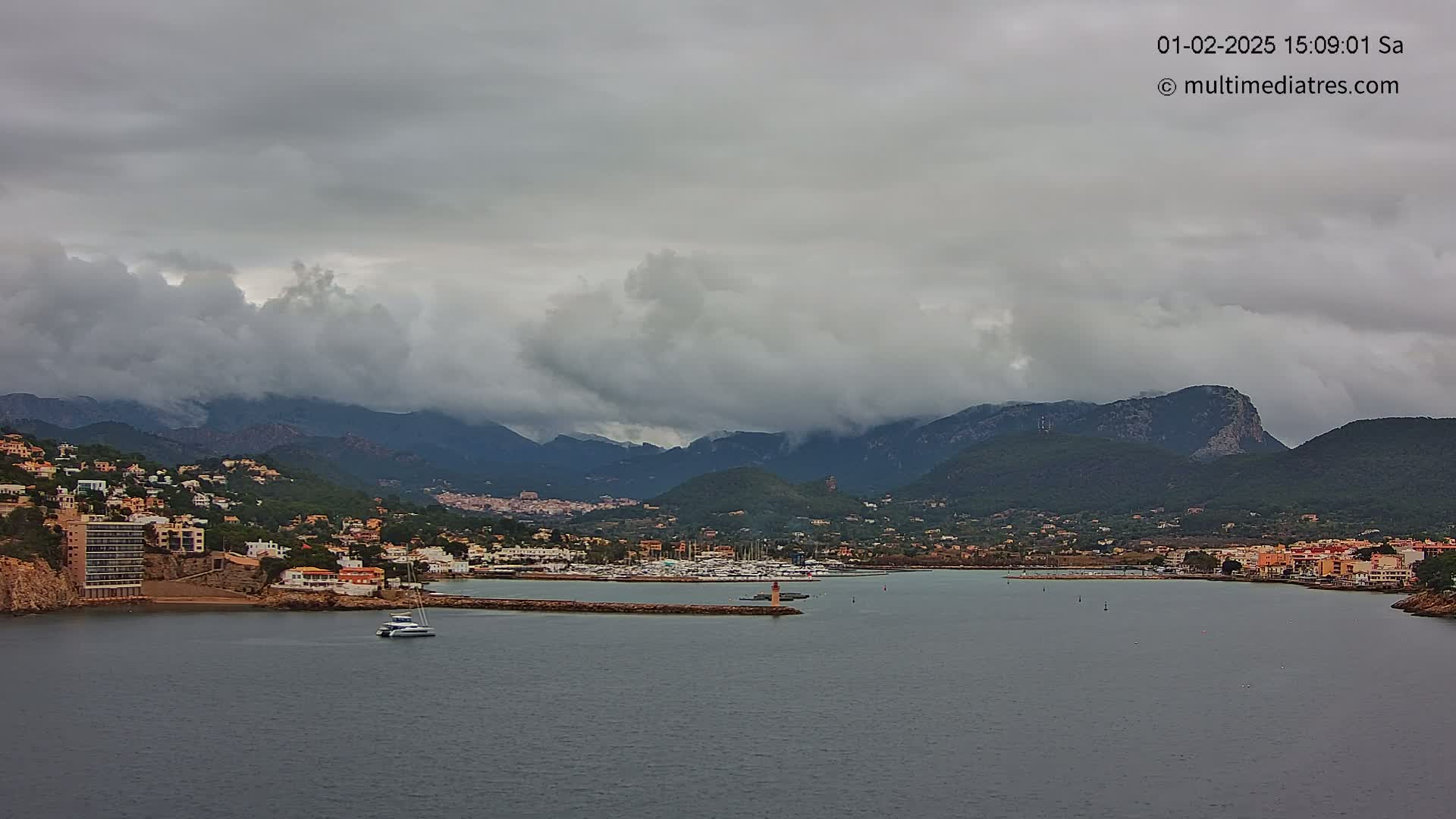A coastal town nestled in a mountain valley under a cloudy sky, with a marina filled with boats visible in the foreground.