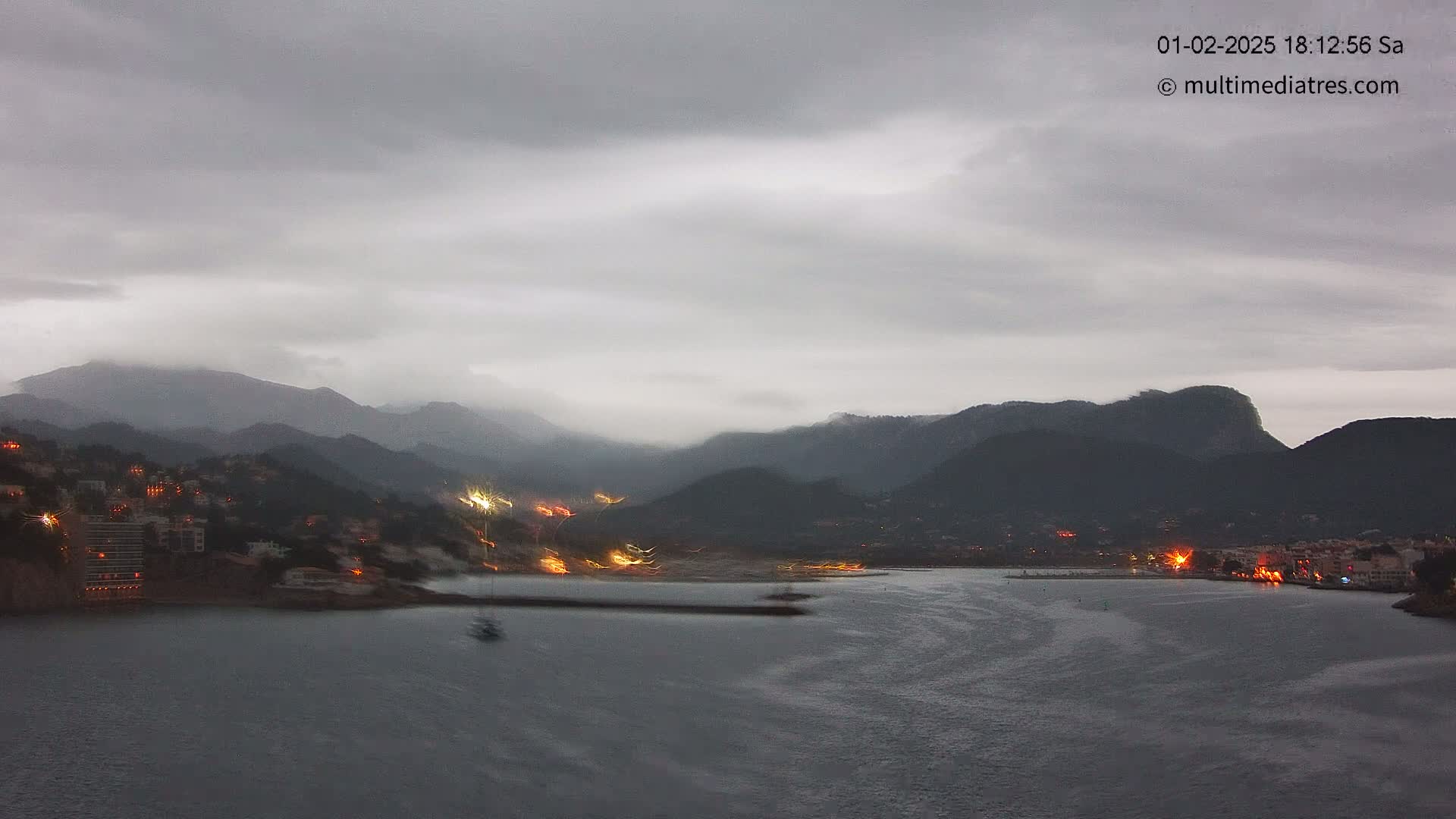 A coastal town at twilight is nestled between dark, mist-covered mountains under a cloudy sky.