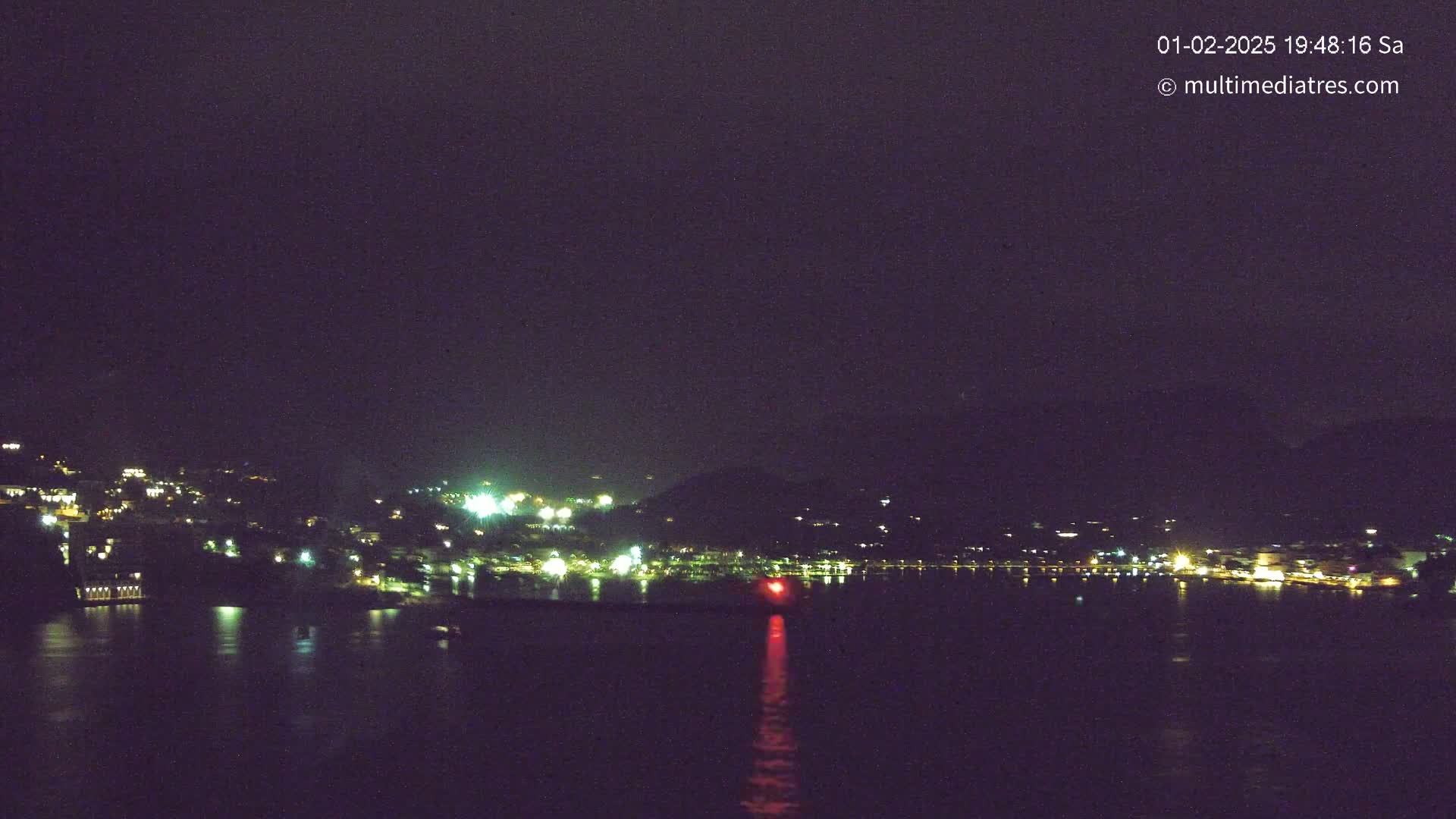 A nighttime view of a town situated on a hillside overlooking a body of water, with lights illuminating the buildings and a red light visible on the water.