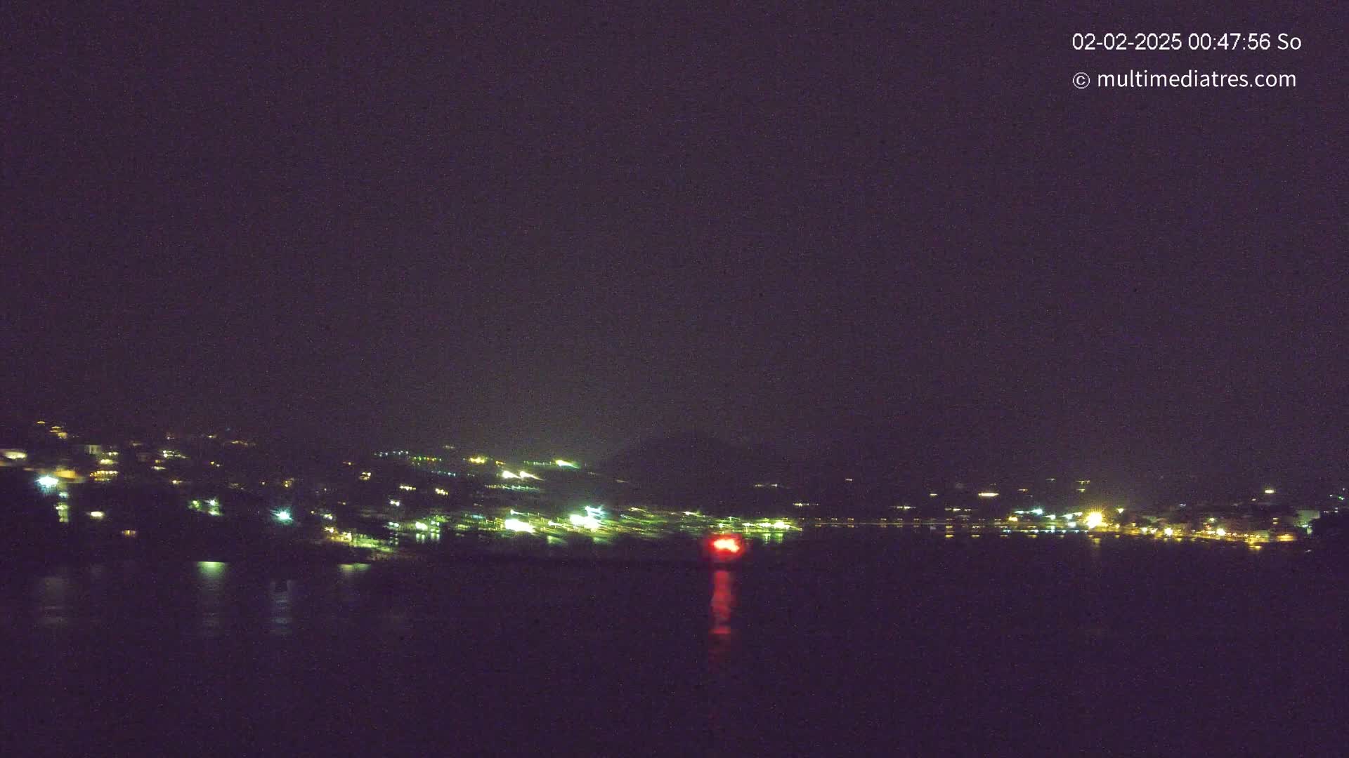 A nighttime view of a town's lights across a dark body of water under a cloudy sky.