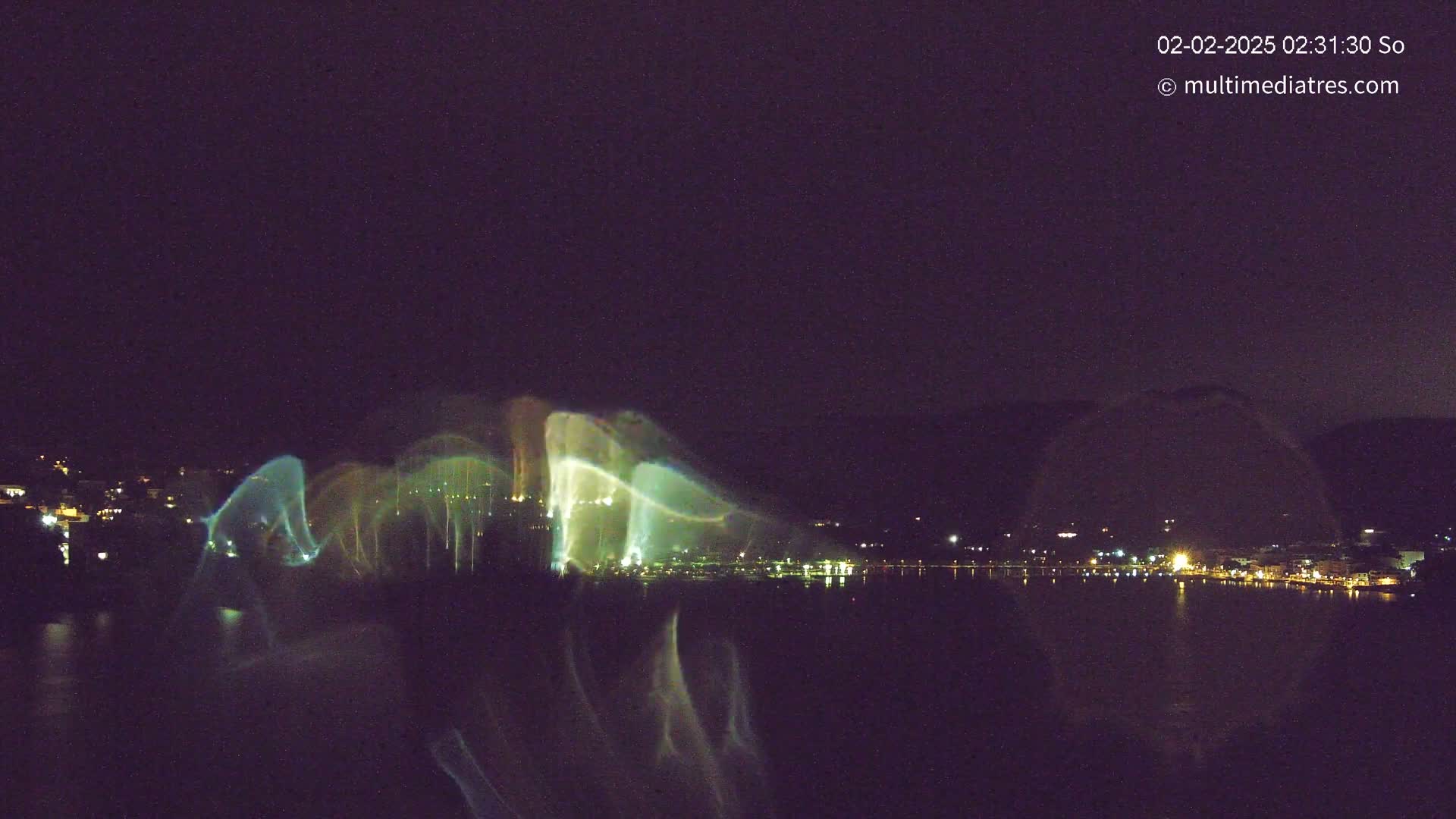 A nighttime long-exposure shot shows a city on a body of water with a light show or water fountain display casting colorful streaks across the dark sky.
