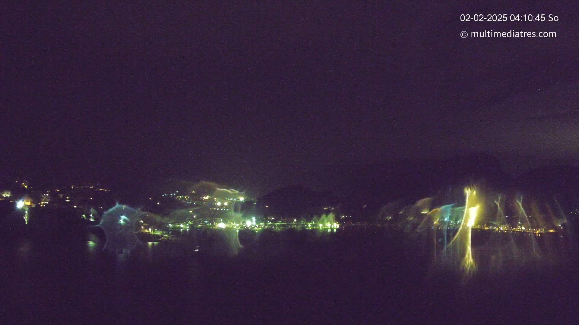 A nighttime long-exposure shot shows a body of water reflecting illuminated hills and a large fountain display.