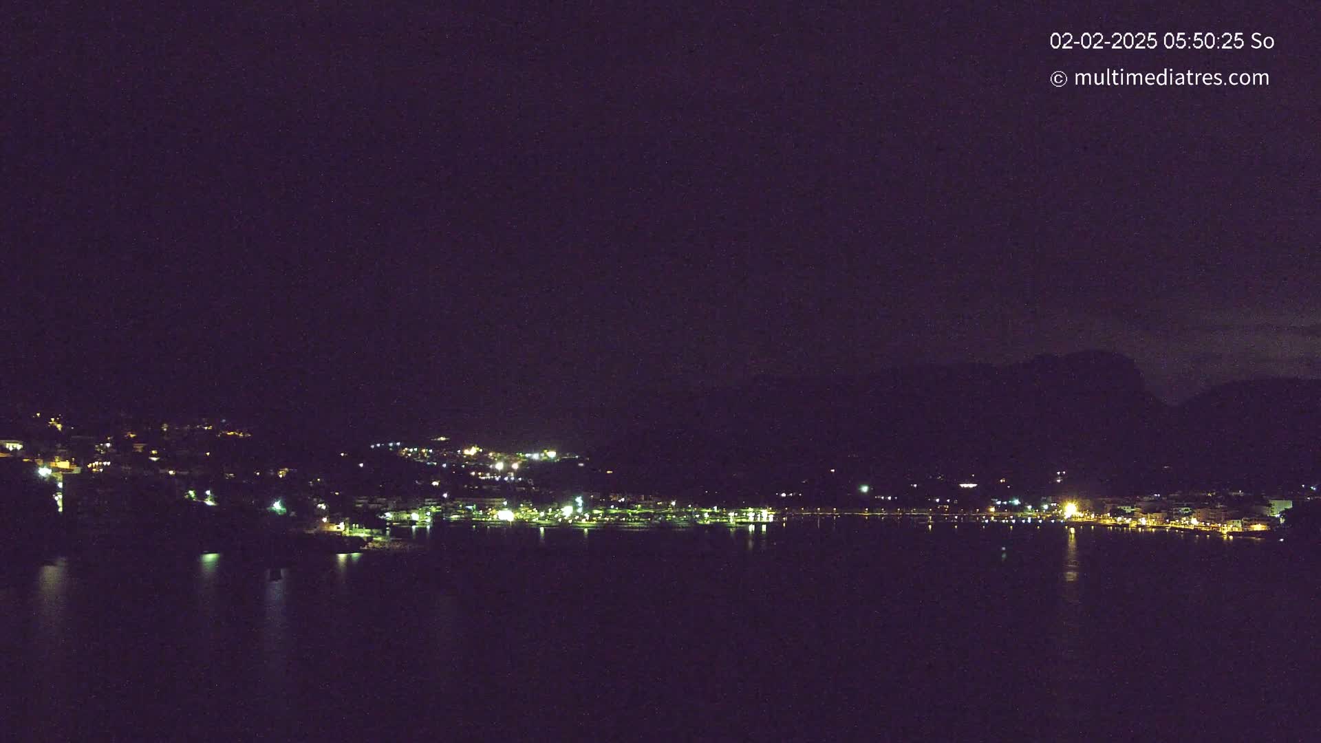 A nighttime long shot shows a town's lights scattered along a dark shoreline and reflected on a calm body of water, with dark hills rising behind it under a clear, dark sky.