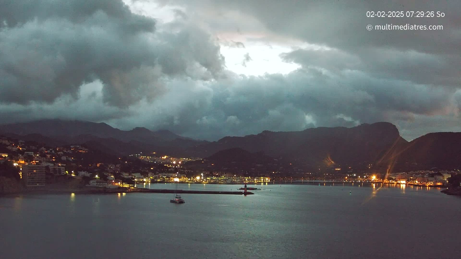 A coastal town at night, illuminated by lights, is nestled between dark mountains under a stormy, overcast sky.