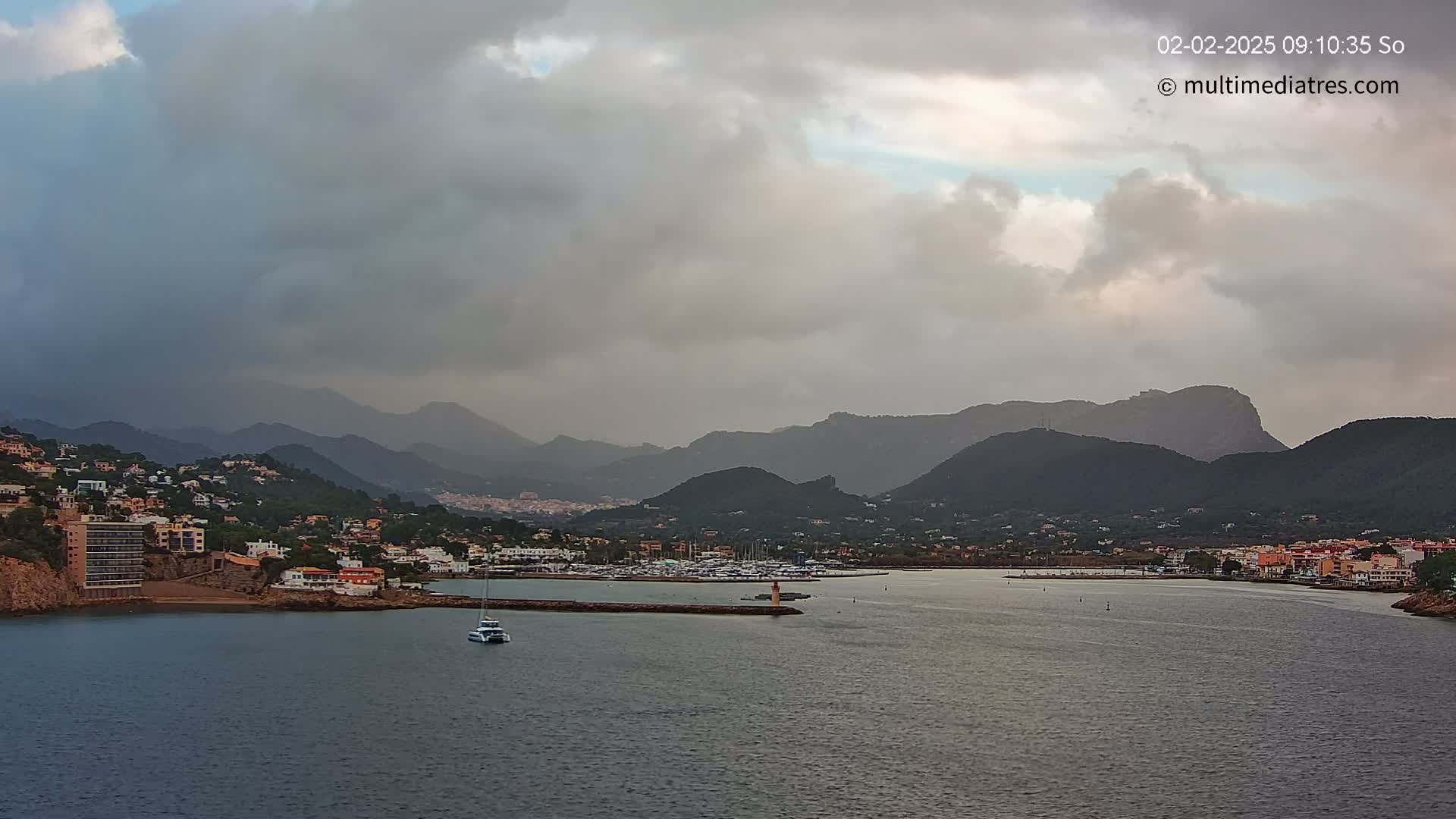 A coastal town nestled between dark mountains under a cloudy sky, with a marina and a small boat visible in the calm water.