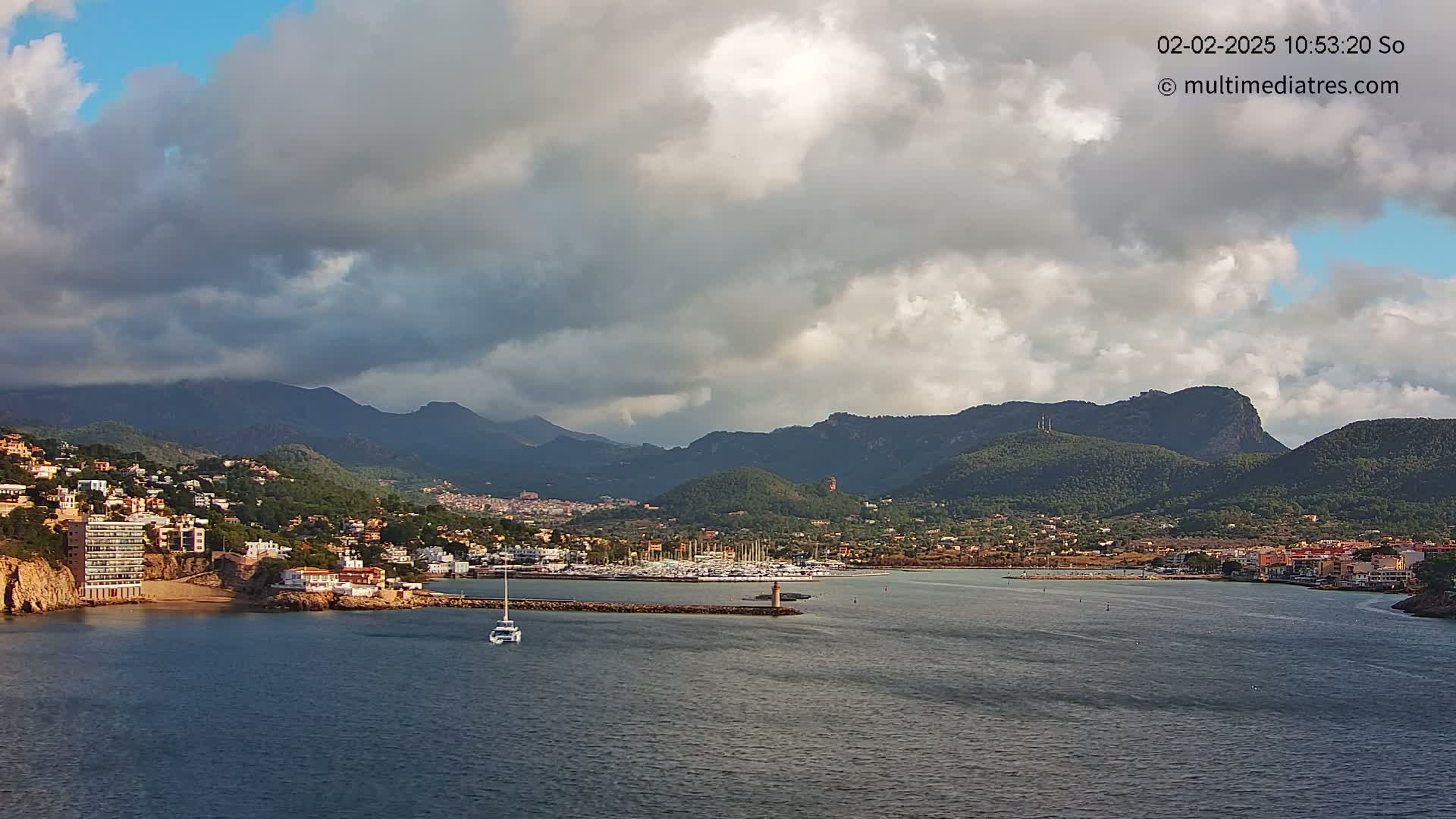 A partly cloudy day shows a coastal town nestled between mountains and a harbor filled with boats, with a sailboat visible in the water.