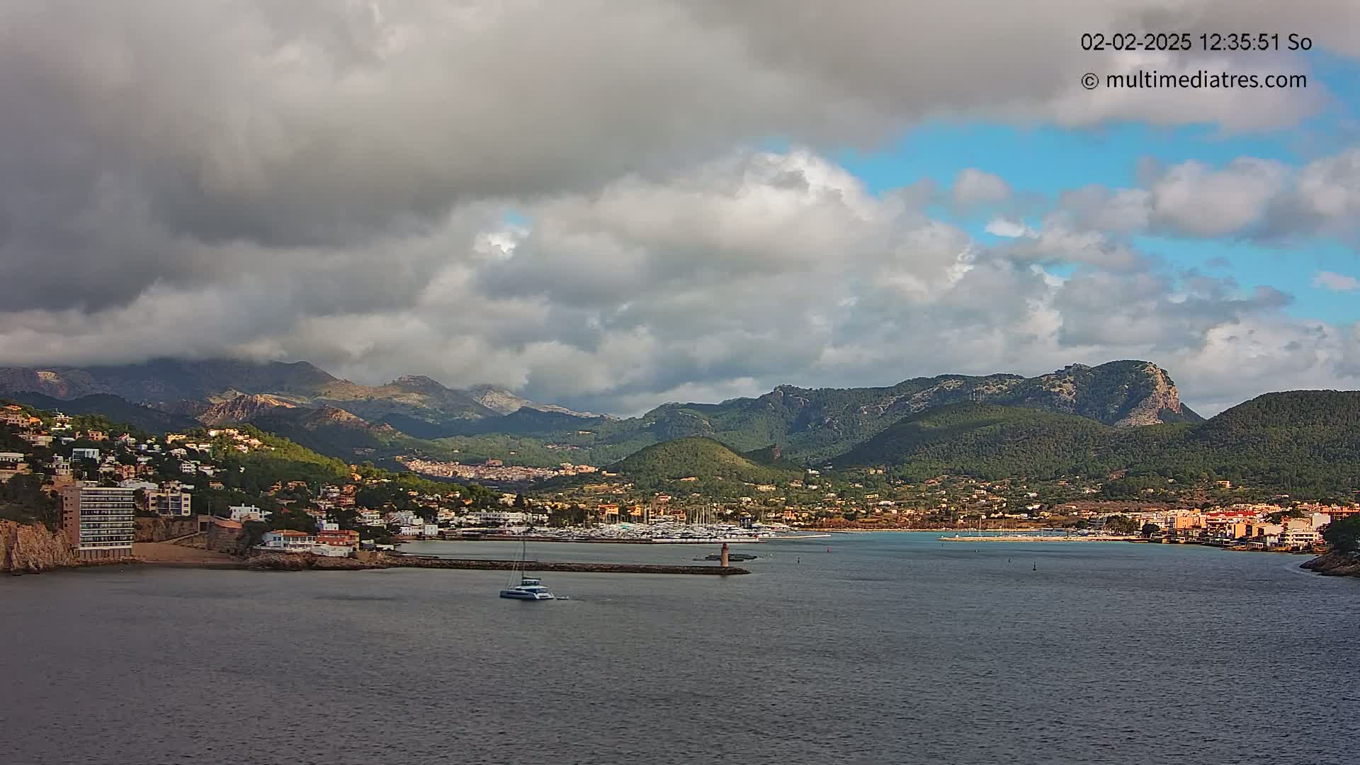 A coastal town nestled between mountains under a partly cloudy sky, with a marina filled with boats and a single sailboat in the bay.