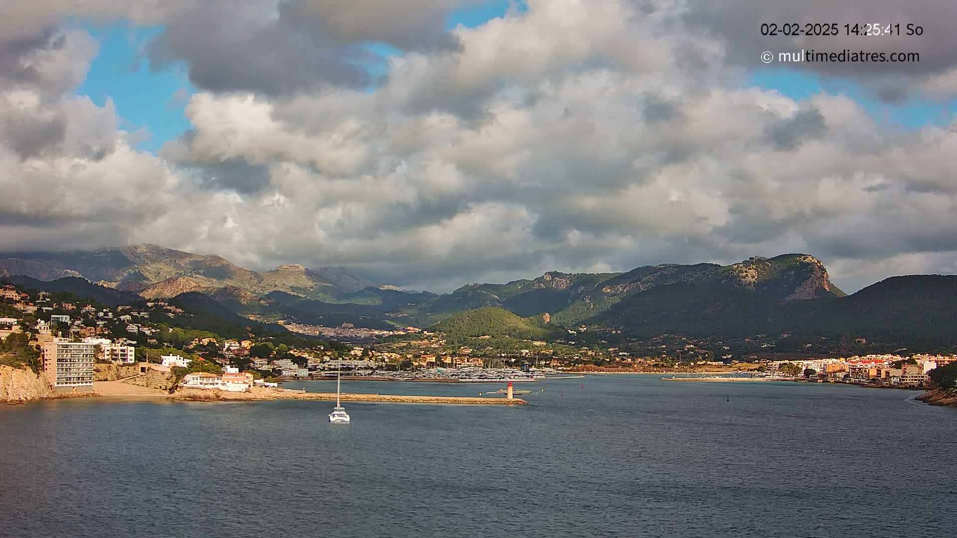 A partly cloudy day shows a coastal town nestled between mountains and a calm body of water, with a small boat visible in the harbor.