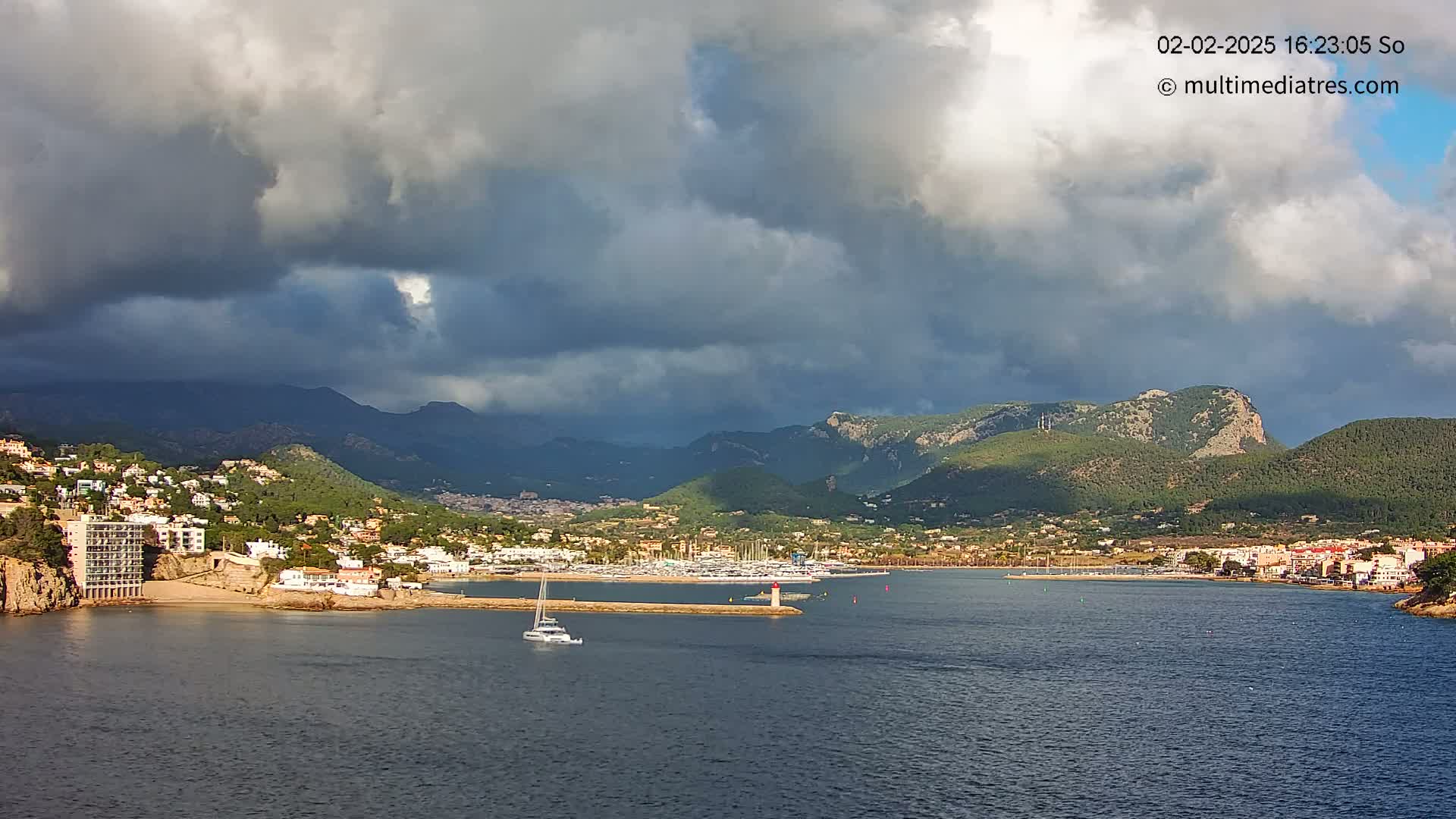 A coastal town nestled in a mountainous region is partially obscured by dark, heavy clouds over a calm body of water.