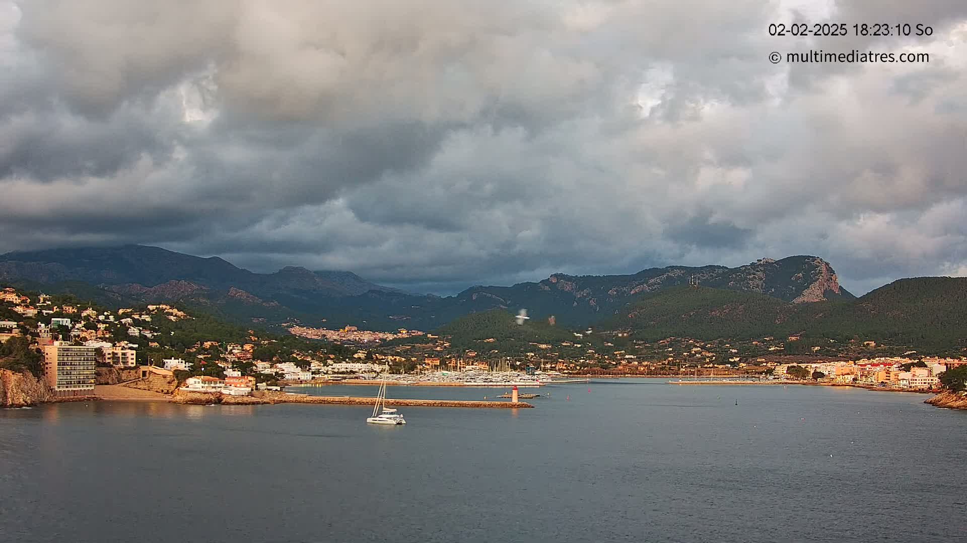 A coastal town nestled against dark, cloudy skies and mountains, features a harbor with several boats, under a partly cloudy sky.