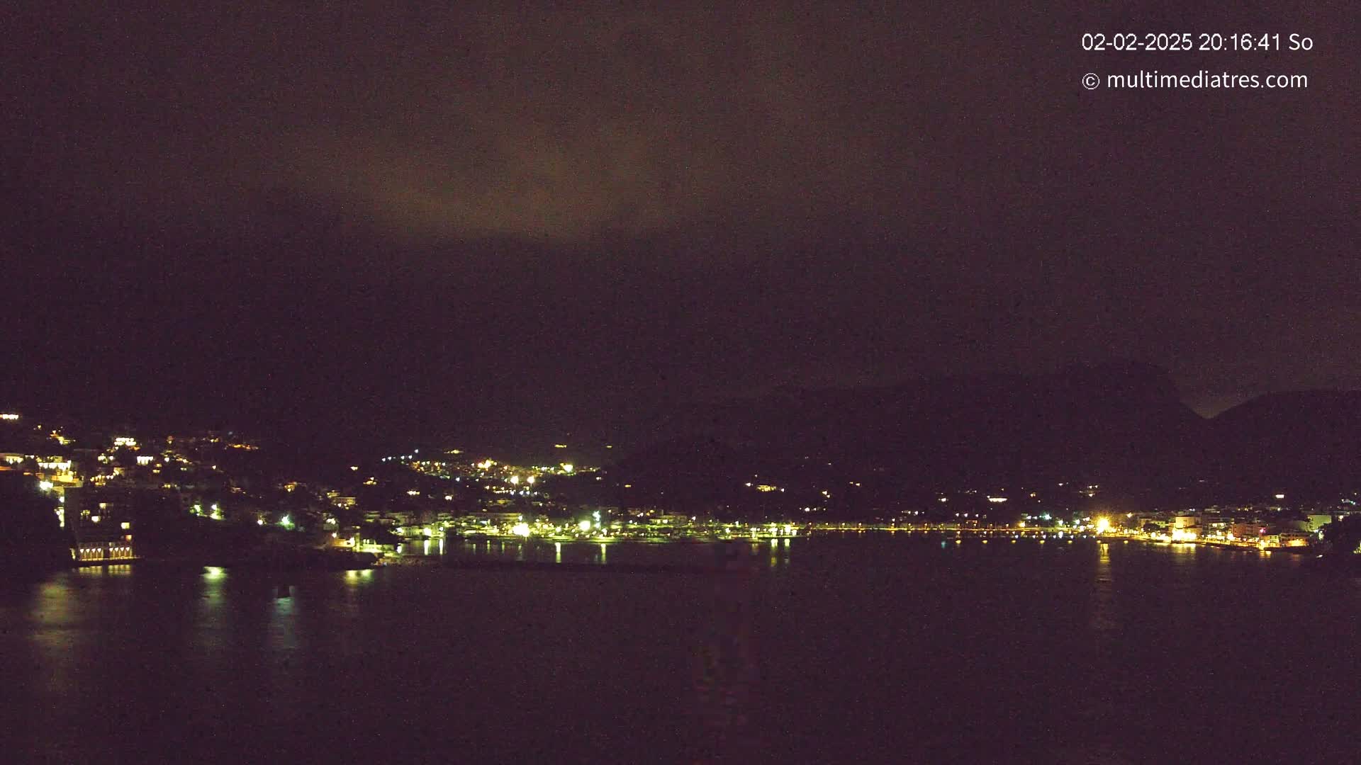 A nighttime view of a coastal town nestled between dark hills, illuminated by scattered lights, under a cloudy sky.