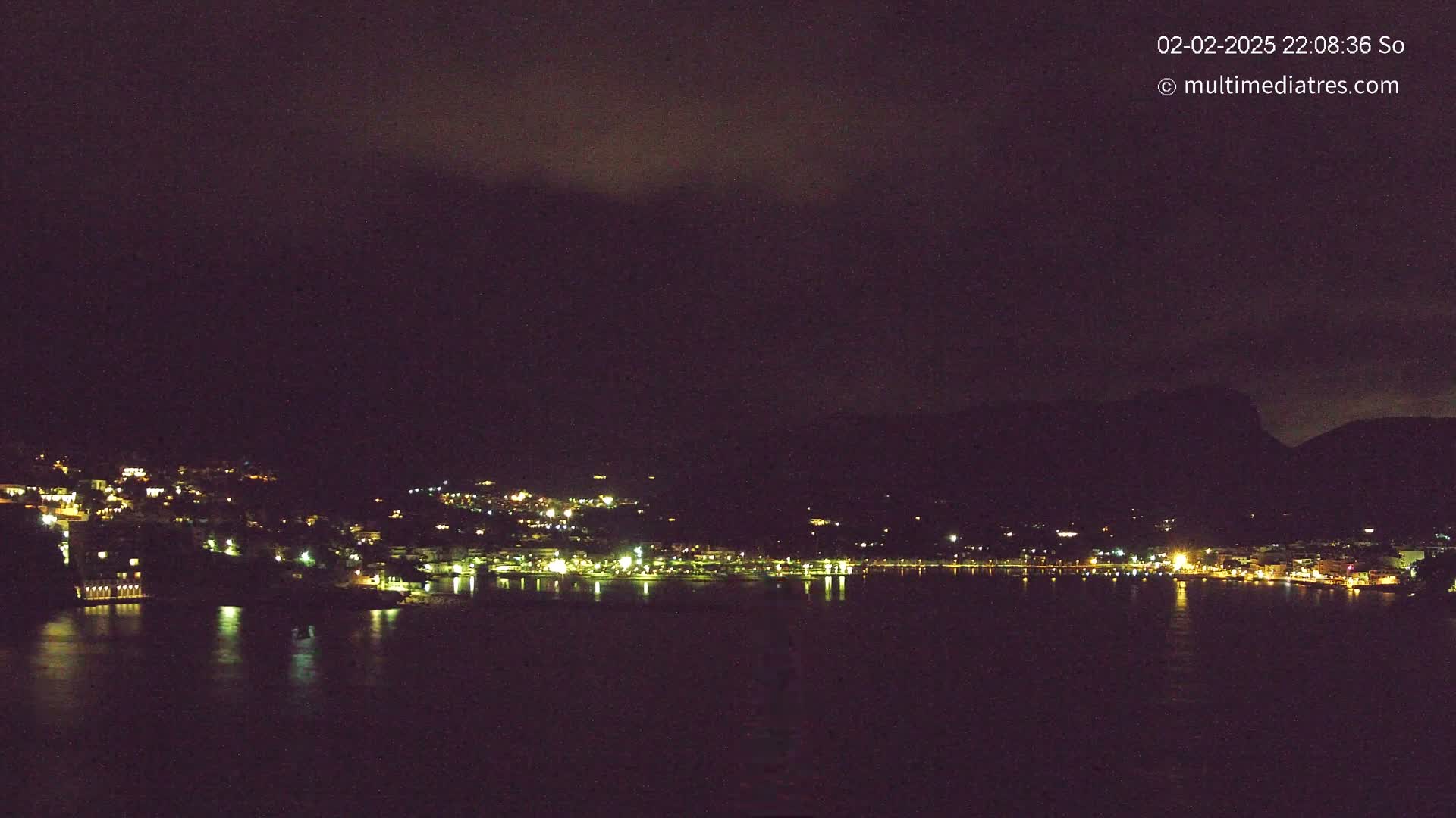 A nighttime view of a town situated along a dark body of water, nestled between hills under a mostly cloudy sky.