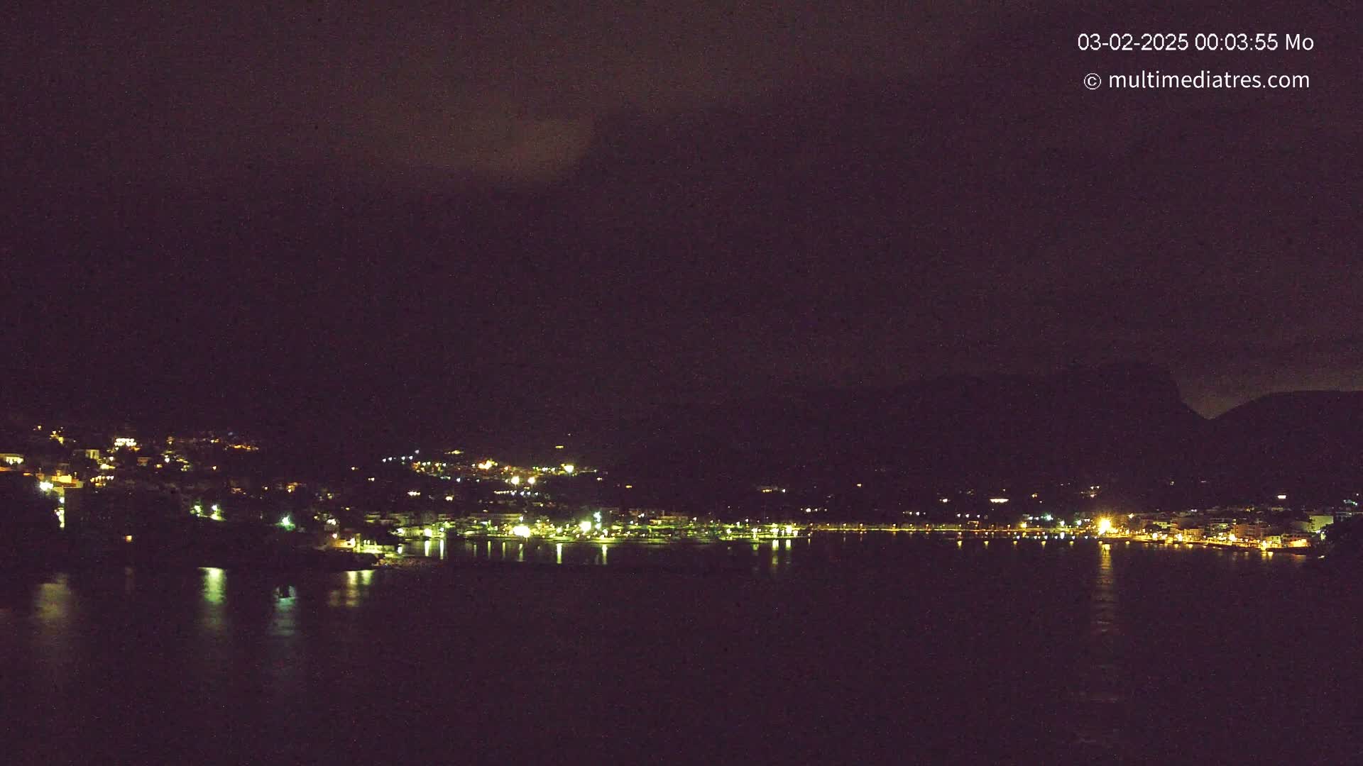 A nighttime view of a coastal town with lights illuminating buildings along a dark body of water under a mostly cloudy sky.