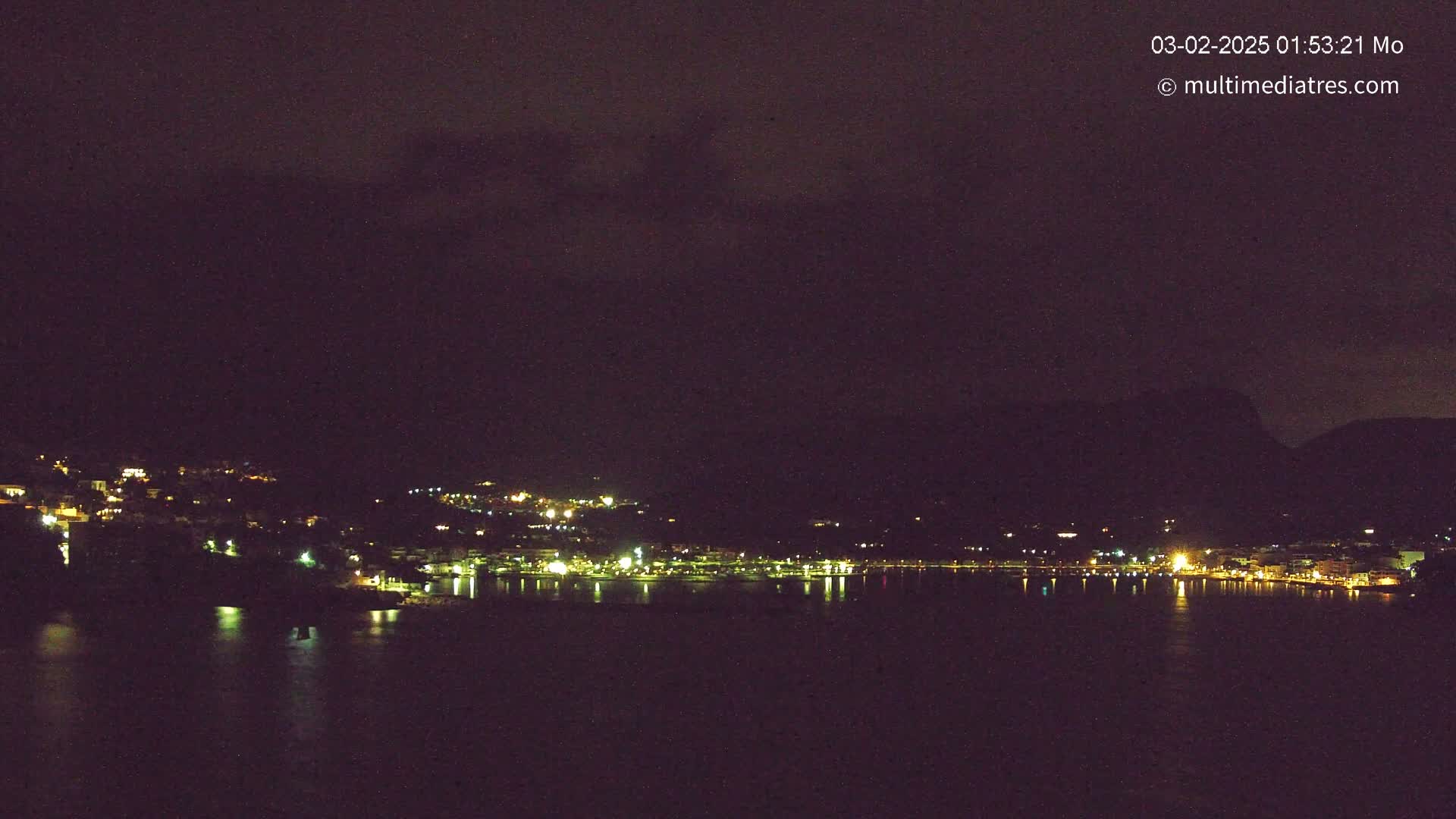 A nighttime view of a coastal town with its lights reflecting on calm water under a dark, cloudy sky.