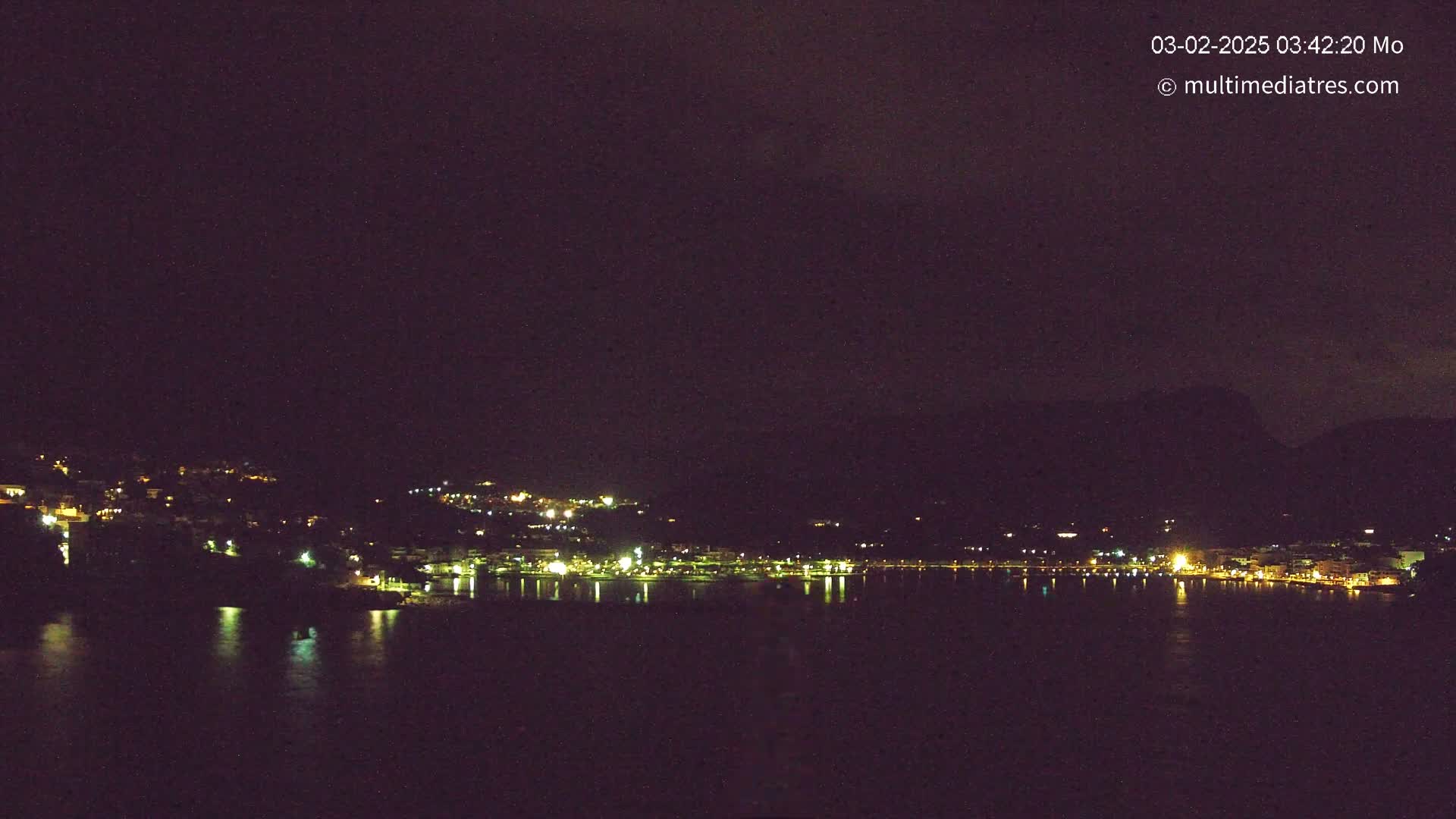 A nighttime view of a town's lights scattered along a dark, calm body of water, nestled against a hillside under a dark, cloudy sky.