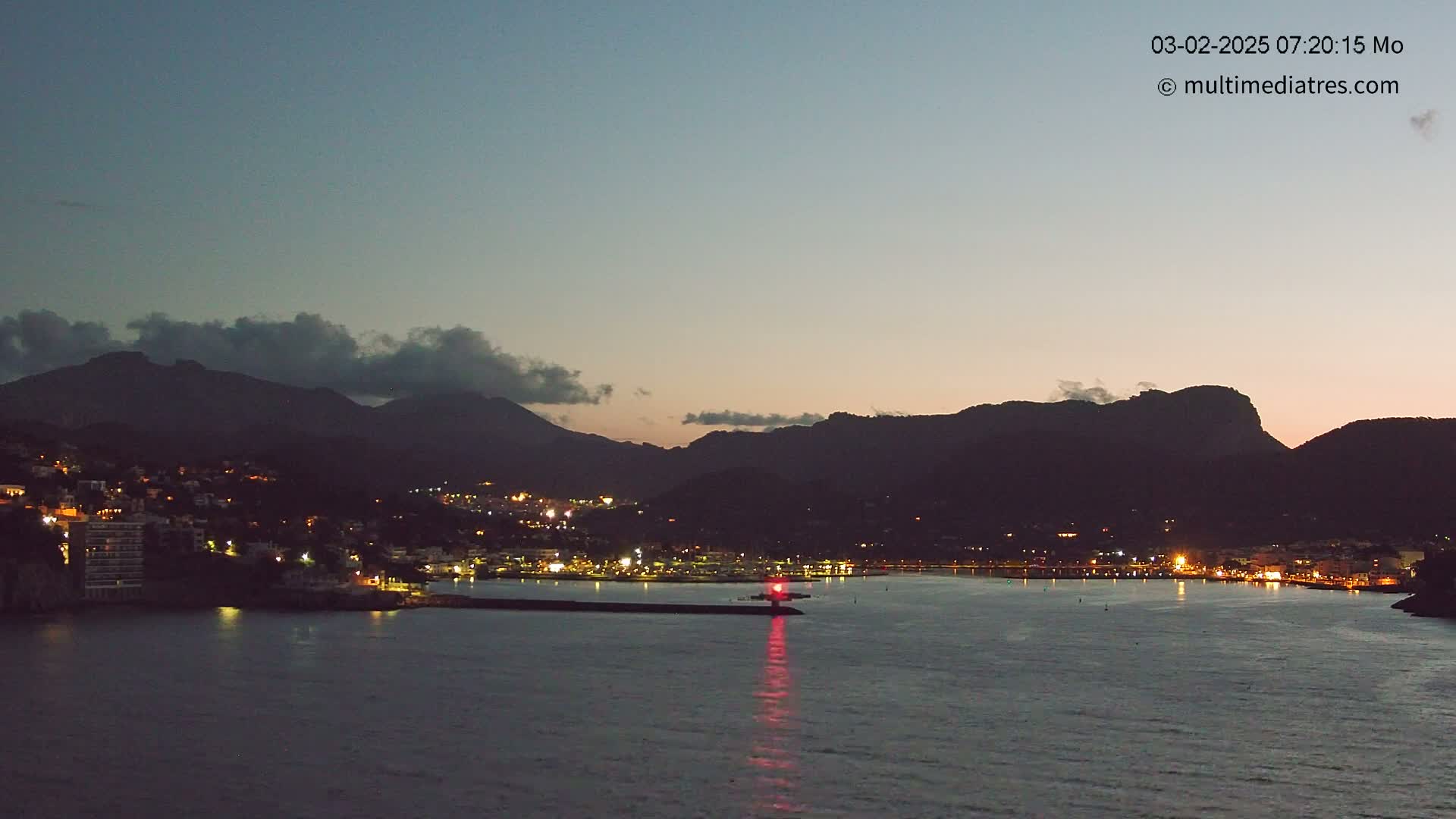 A coastal town at twilight, nestled between dark mountains under a mostly clear sky, with a harbor illuminated by lights.