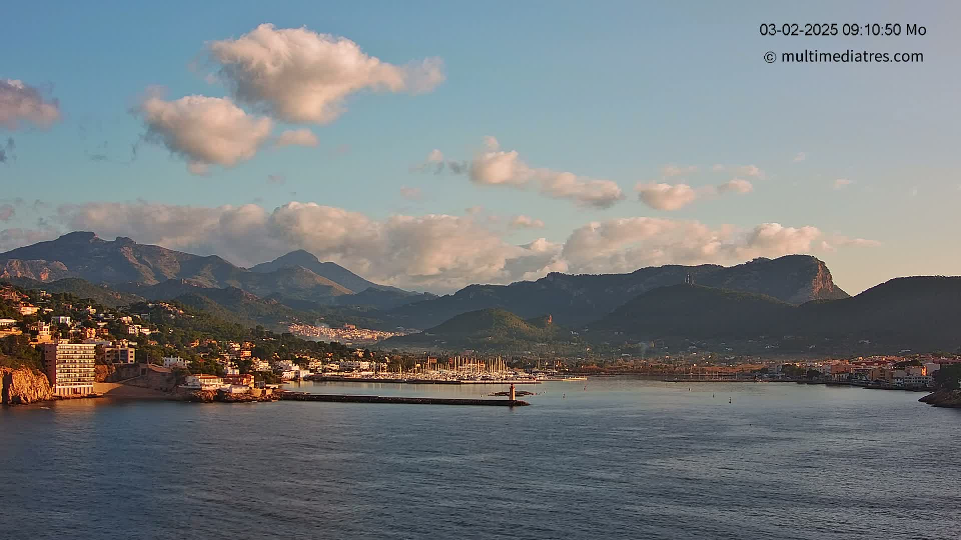 A coastal town nestled at the base of mountains, with a harbor full of boats, under a partly cloudy sky.