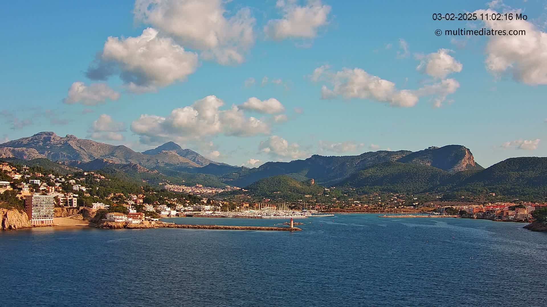 A coastal town nestled between mountains, with a marina filled with boats, under a partly cloudy sky.