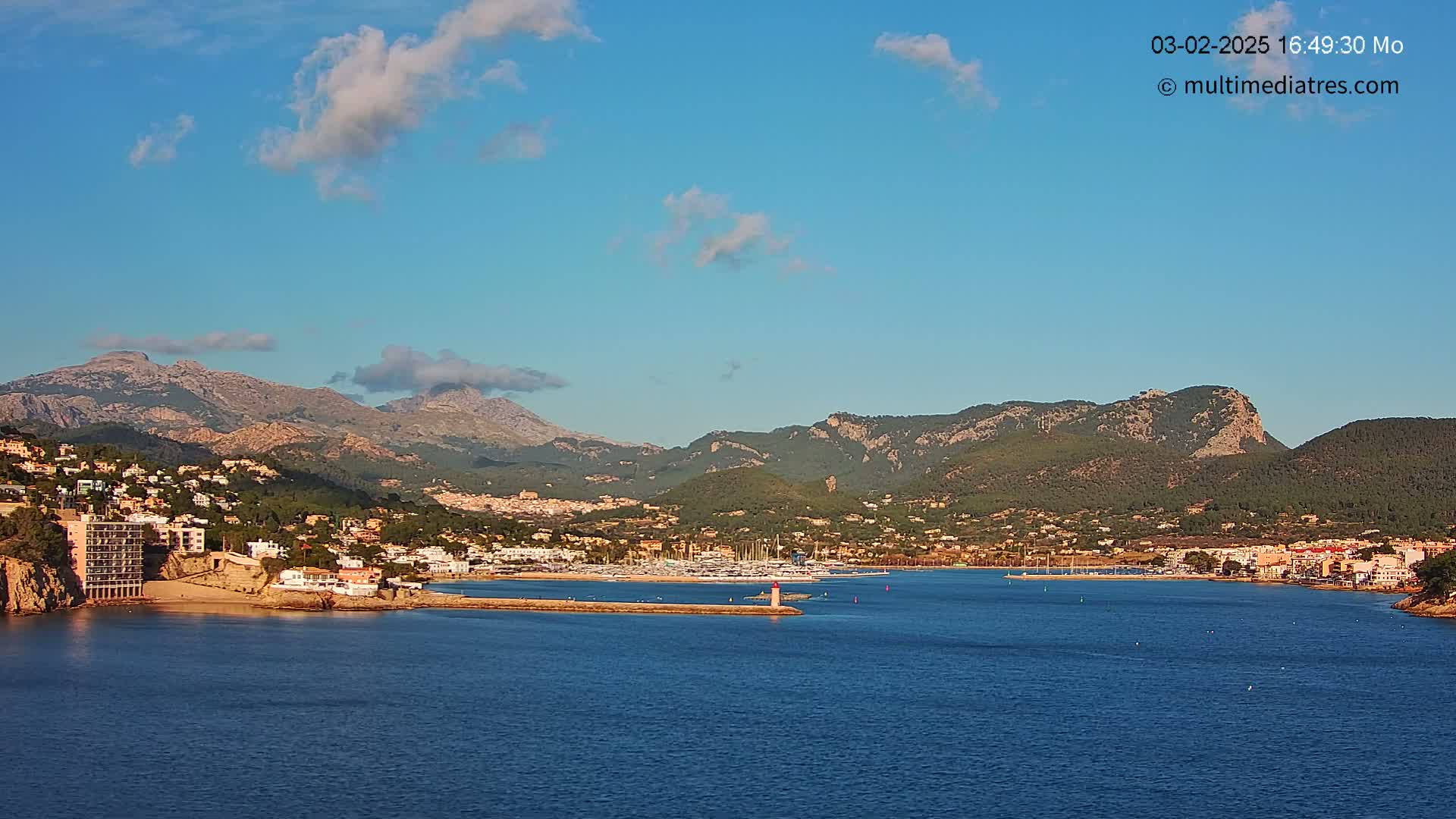 A coastal town nestled between mountains and a calm blue bay under a mostly clear sky with a few clouds.