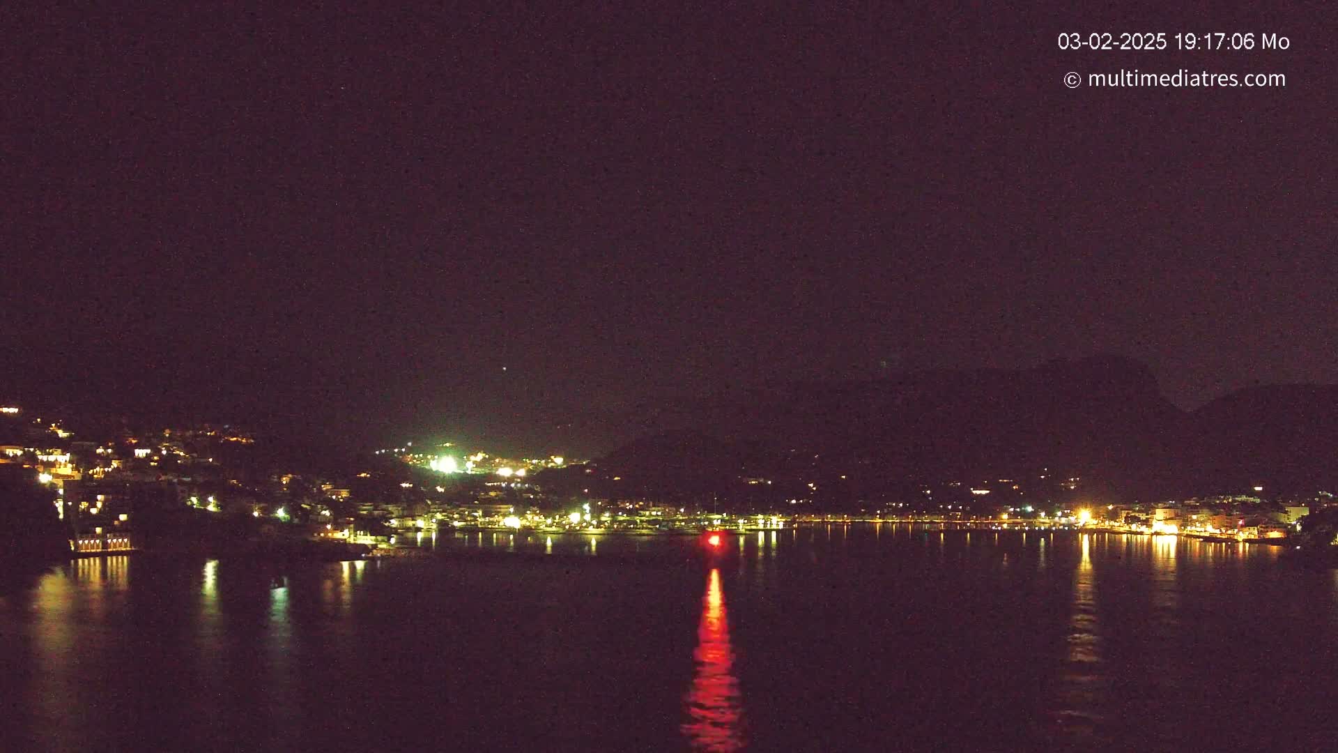 A nighttime view of a town's lights lining a dark shoreline and reflecting on a calm body of water, with a hill in the background under a clear, dark sky.