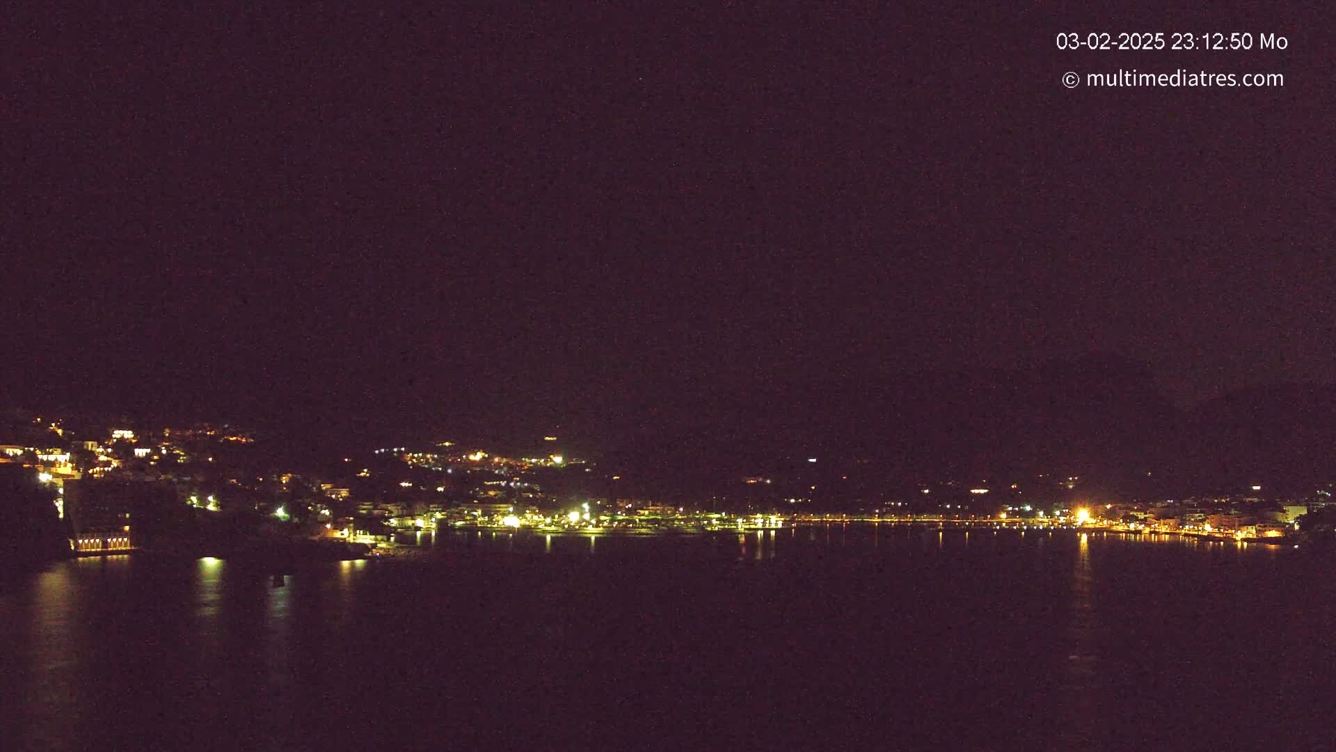A nighttime view of a coastal town with lights illuminating buildings along a dark body of water under a clear, dark sky.