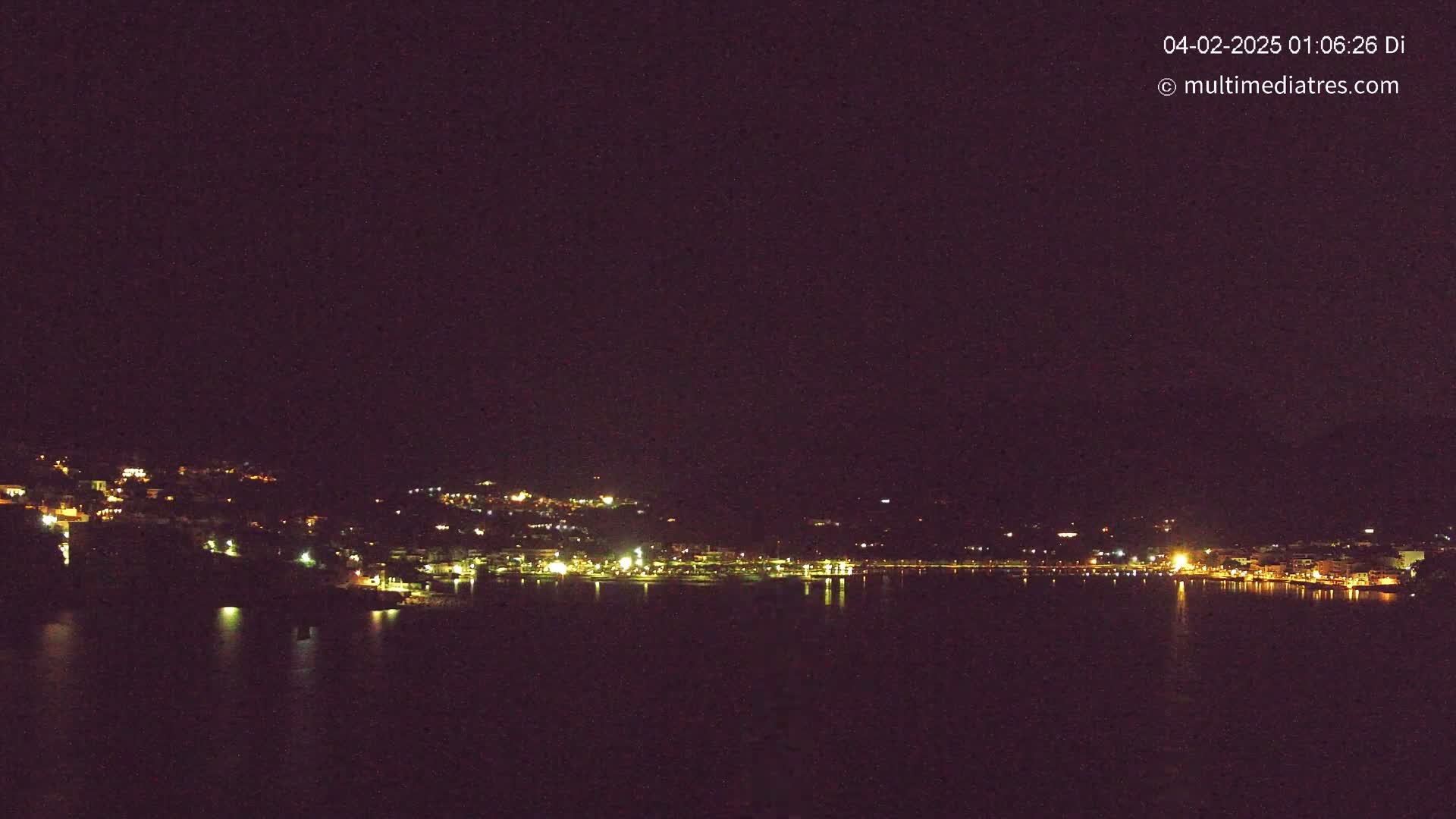A nighttime view of a coastal town with its lights reflecting on a calm body of water under a dark sky.