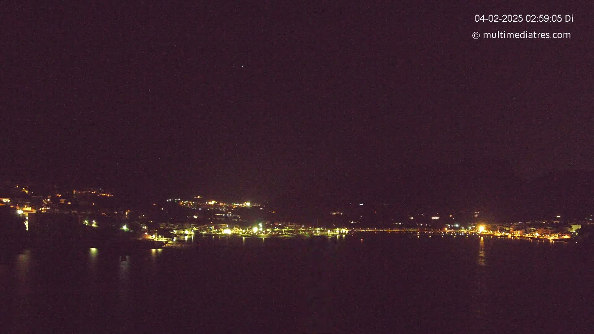 A nighttime view of a coastal town's lights spread across a hillside and reflected in a dark body of water under a clear, starlit sky.