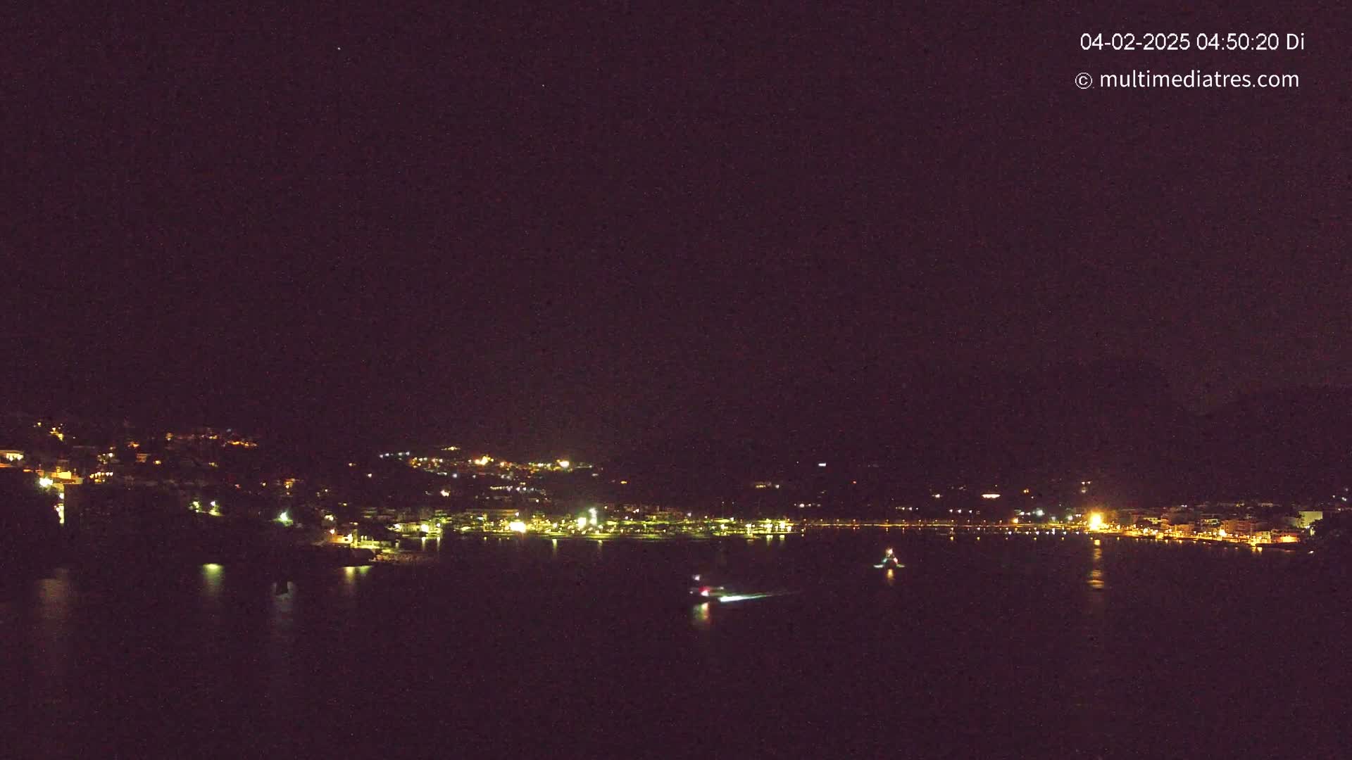 A nighttime, long-range view shows a coastal town with its lights reflecting on dark water, under a clear, star-filled sky.