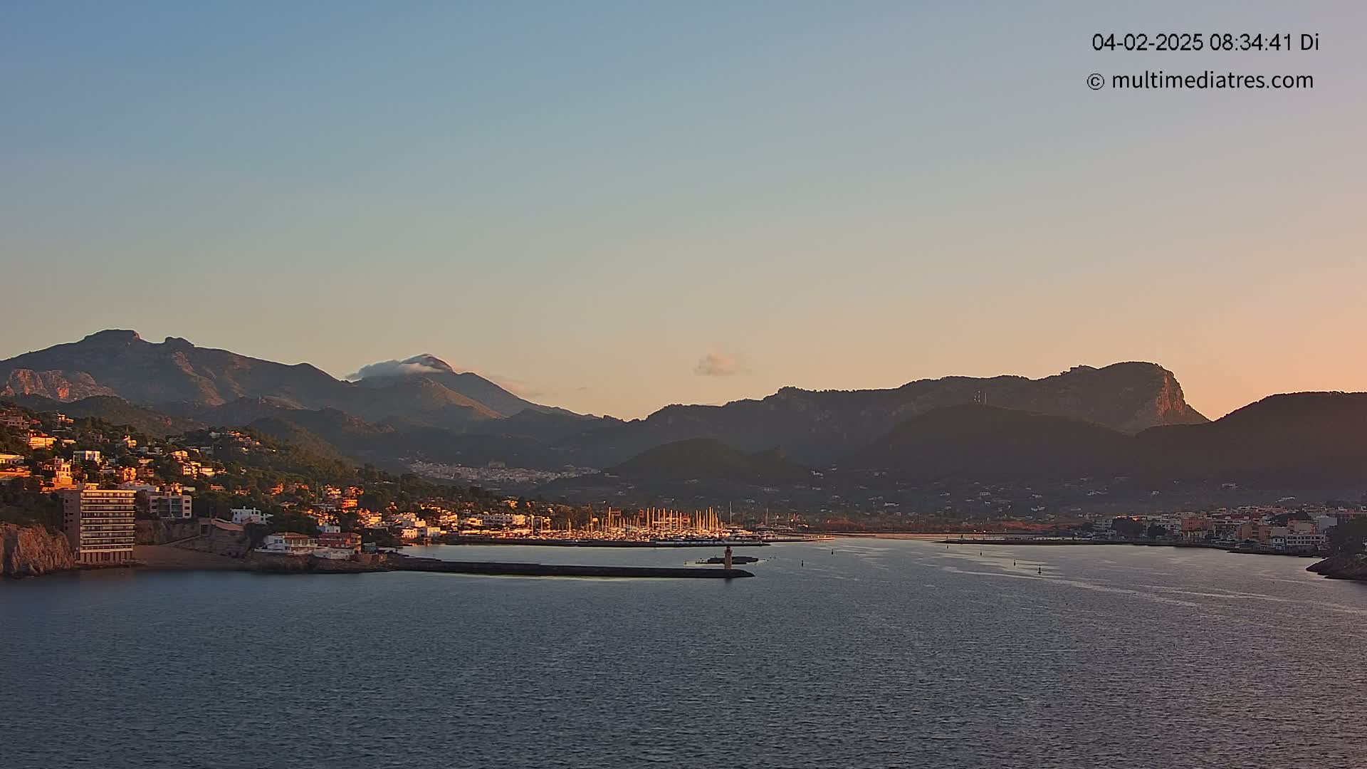 A coastal town nestled against mountains at sunrise, with a calm harbor filled with sailboats.