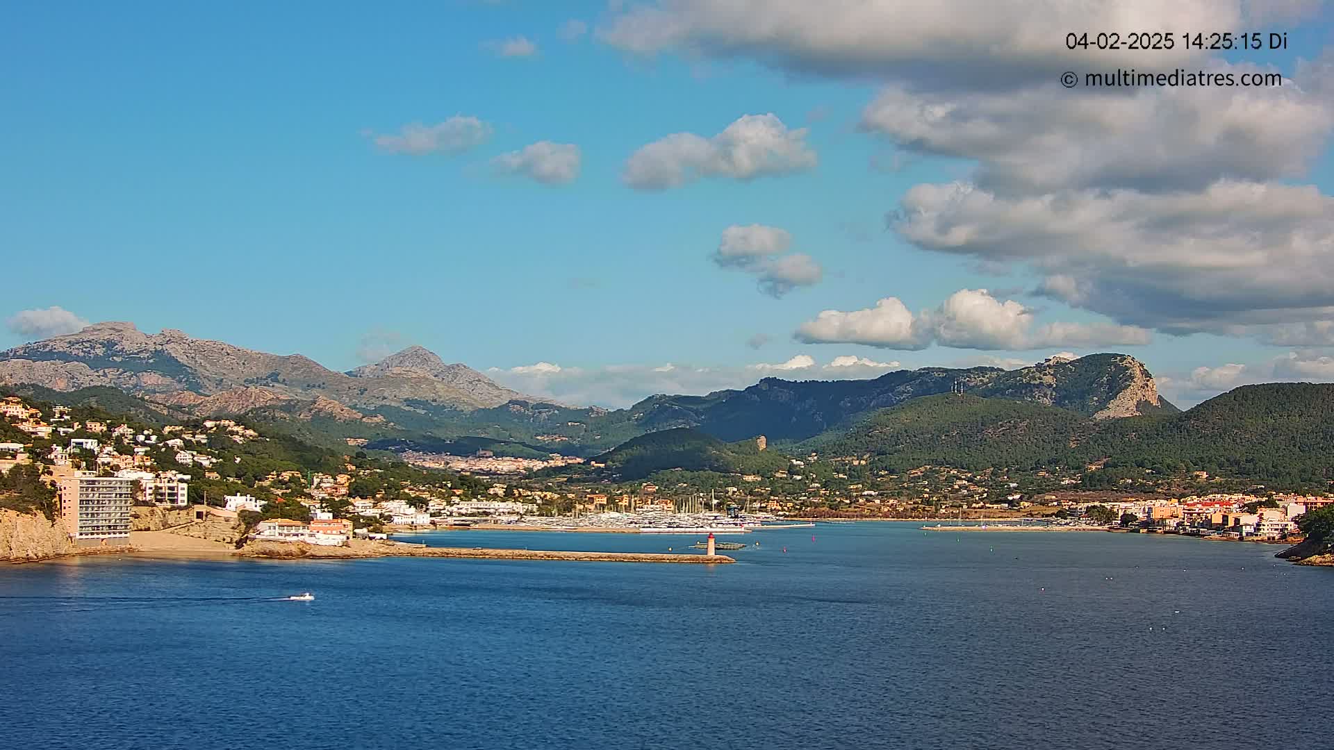 A partly cloudy blue sky overlooks a coastal town nestled between mountains and a calm bay with a marina.