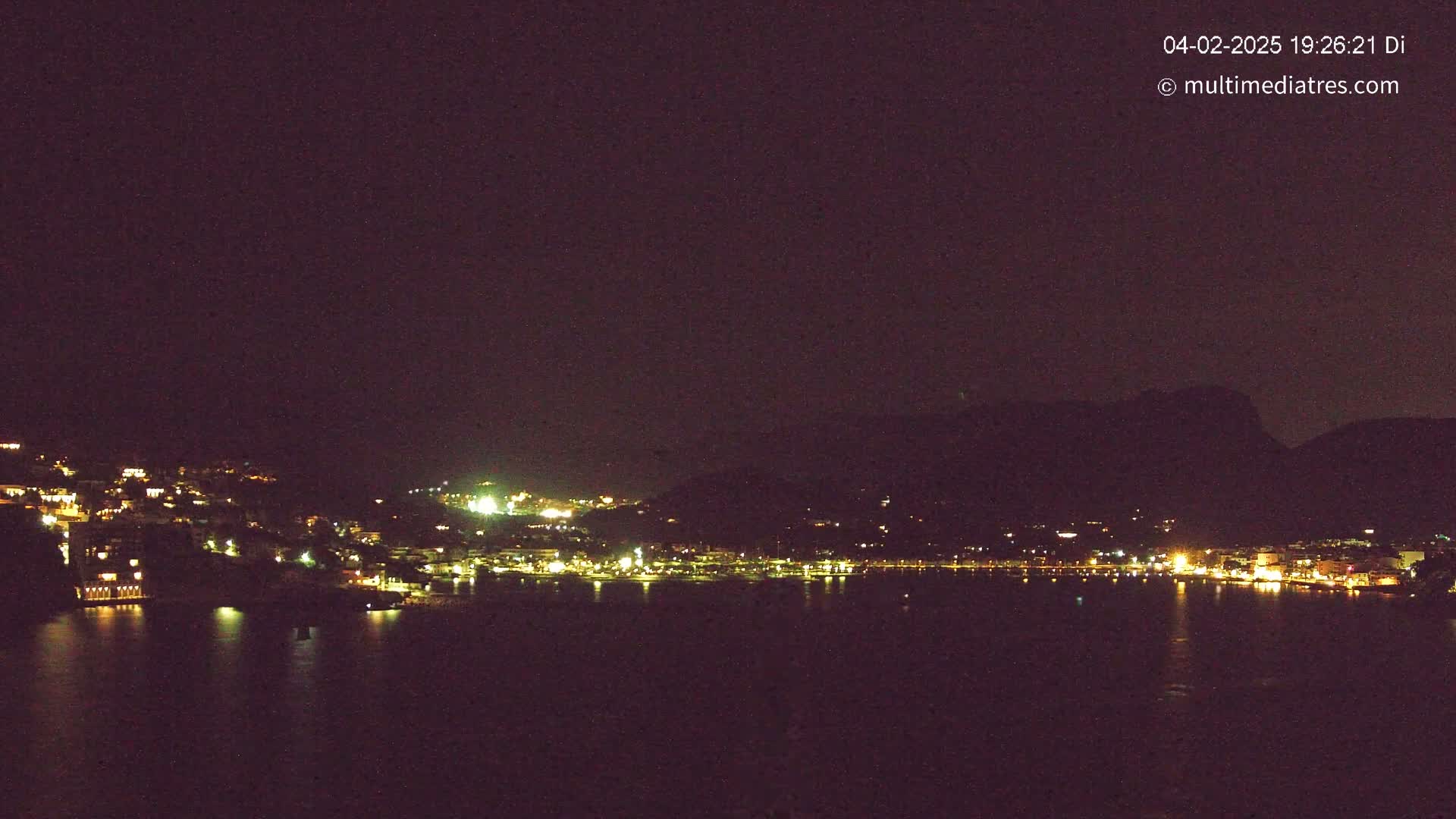 A nighttime view of a town nestled against a dark mountain, illuminated by scattered lights reflecting on a calm body of water.