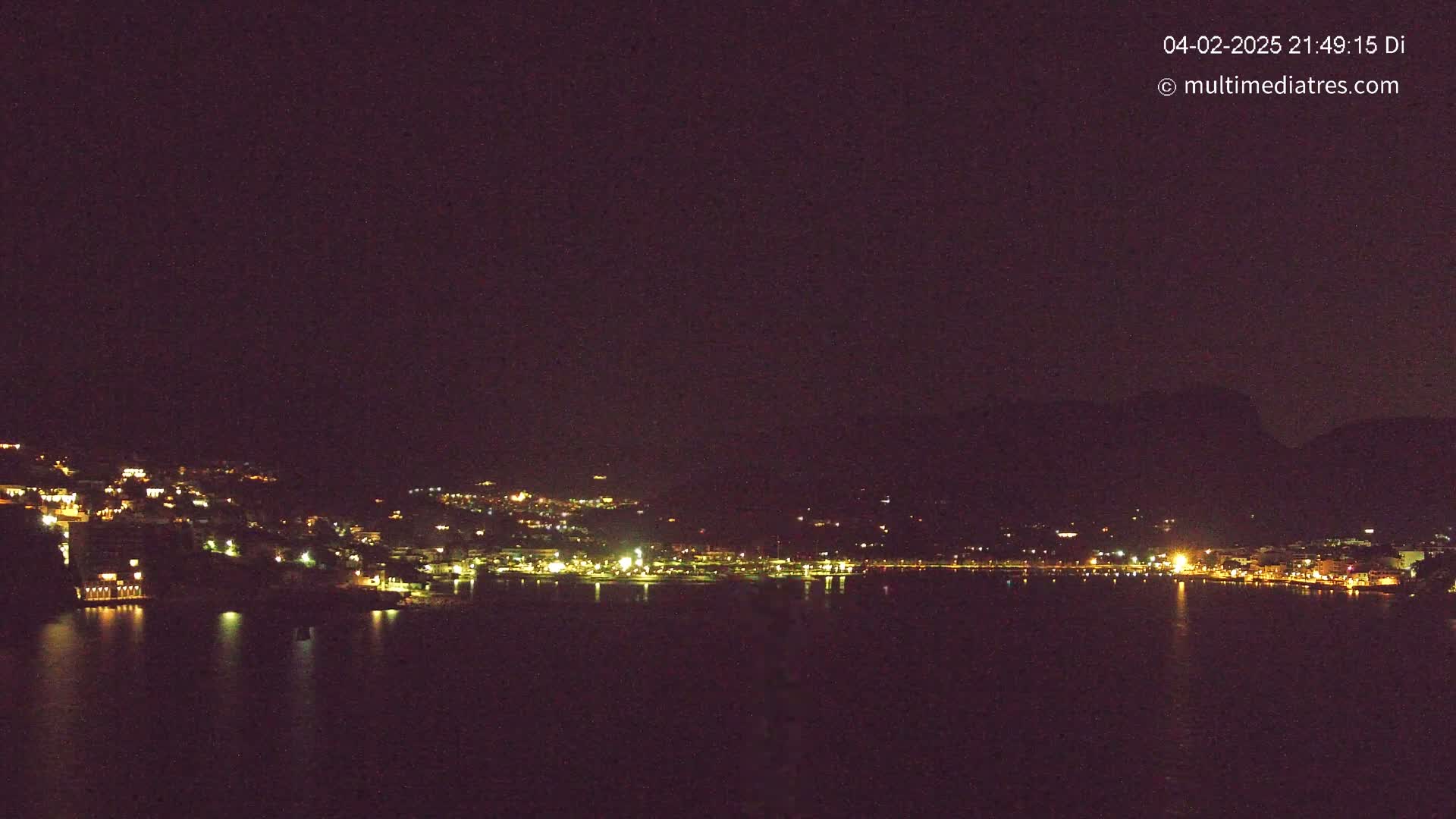 A nighttime view of a town's lights lining a dark body of water, nestled against hills under a clear, dark sky.