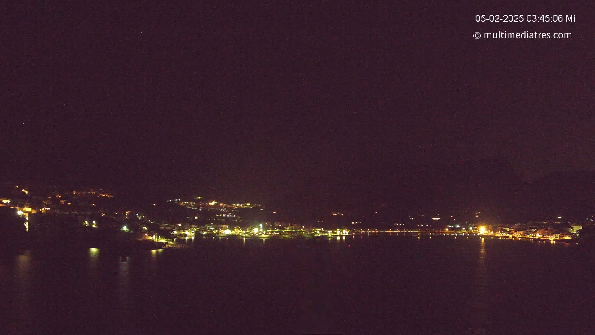 A nighttime view of a town's lights along a dark body of water under a clear, starless sky.