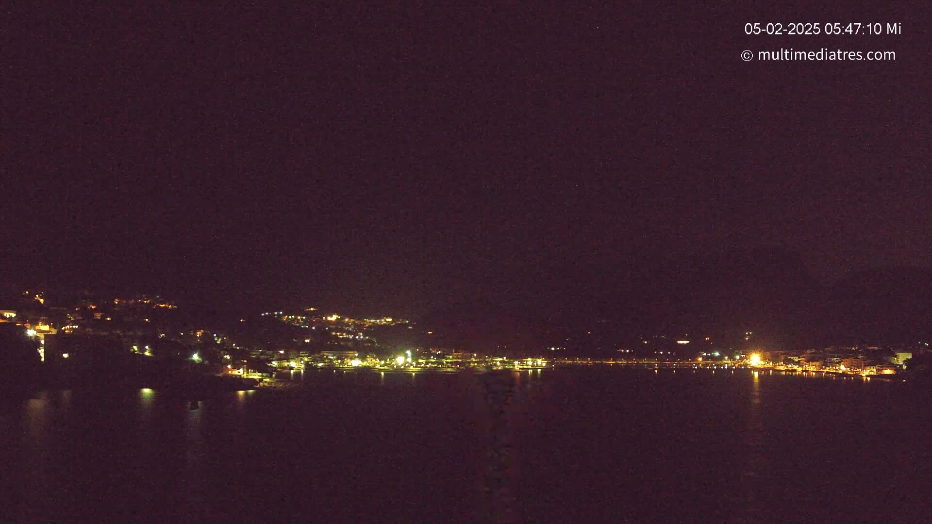 A nighttime view of a town illuminated by lights along a dark body of water under a clear, dark sky.
