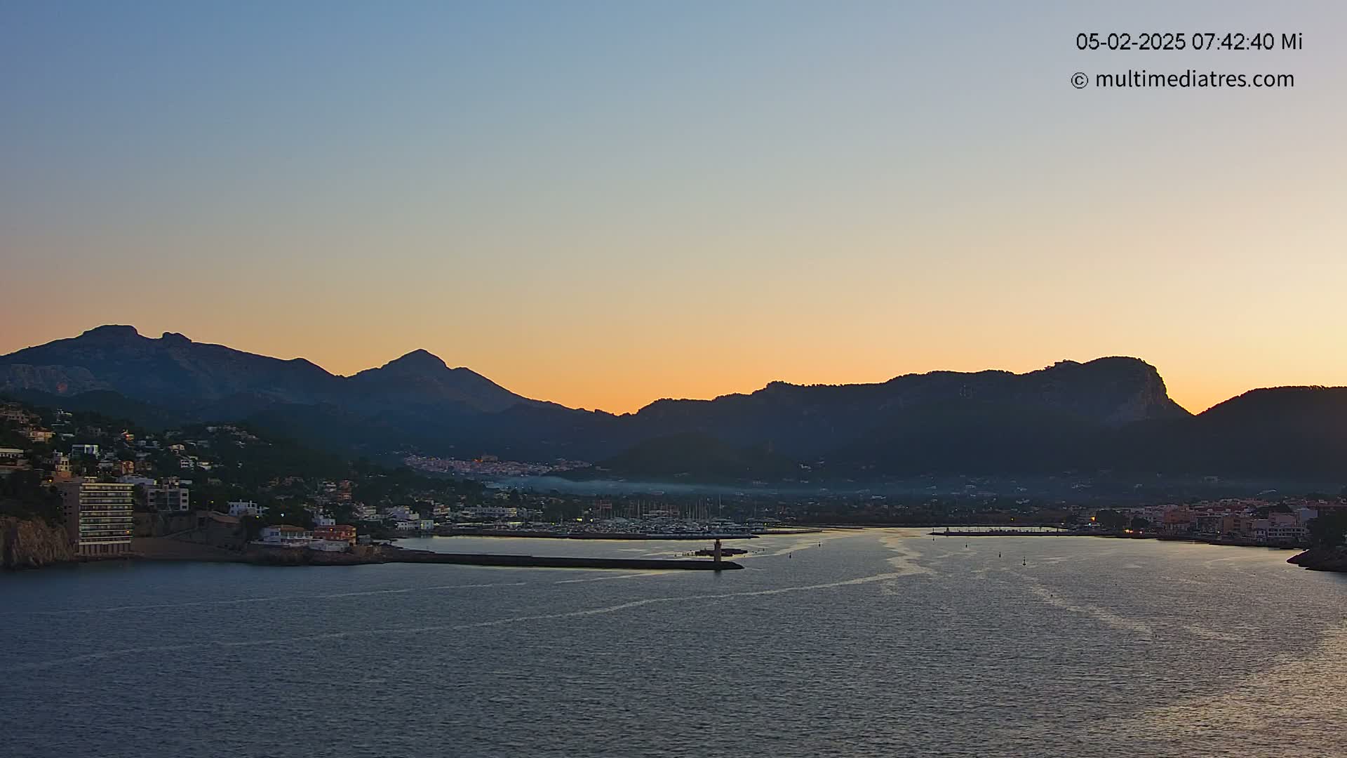 A coastal town nestled at the base of silhouetted mountains is bathed in the warm glow of a sunrise over calm water.