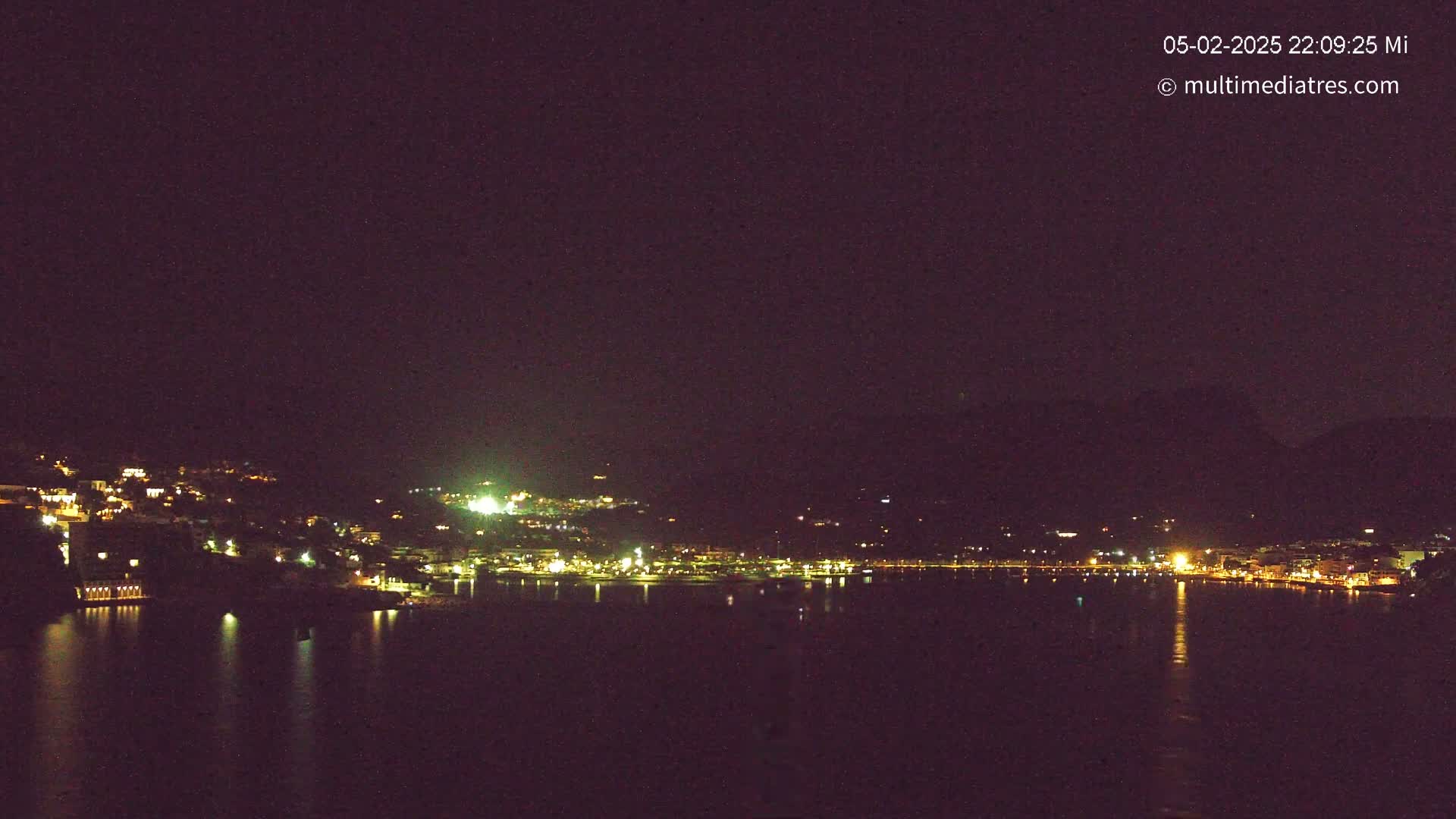 A nighttime view of a coastal town illuminated by lights, reflecting on a calm body of water under a dark sky.