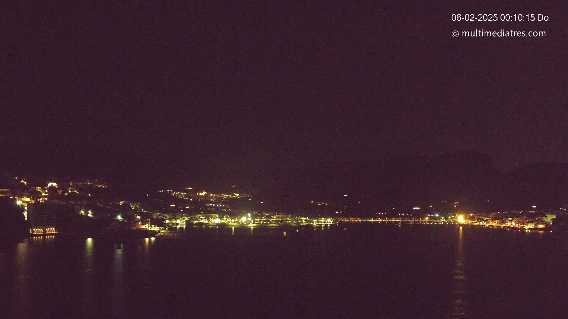 A nighttime view of a coastal town illuminated by lights, overlooking a dark body of water under a clear, dark sky.
